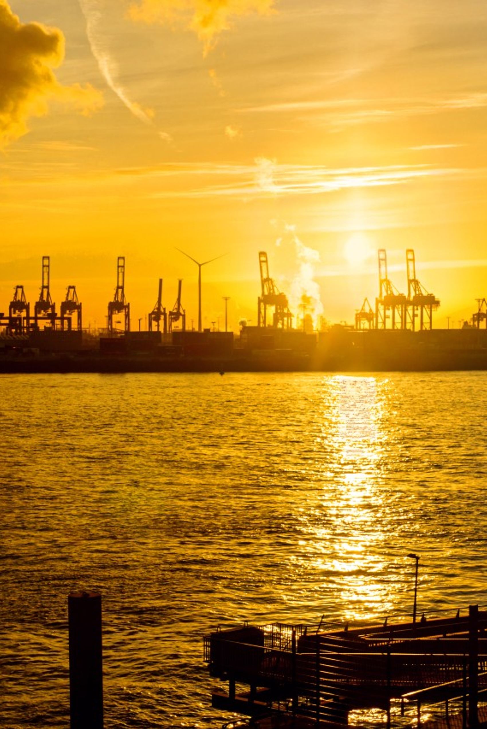 Sonnenuntergang über einem Industriehafen mit Kränen und Windrädern, die am Ufer stehen, reflektiert goldenes Licht auf dem ruhigen Wasser.