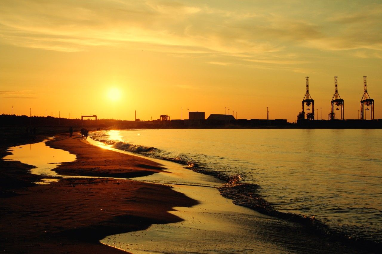 Sonnenuntergang über einem ruhigen Strand mit sanften Wellen, im Hintergrund Industriekräne und Gebäude, die eine harmonische Verbindung von Natur und urbanem Hafen zeigen.