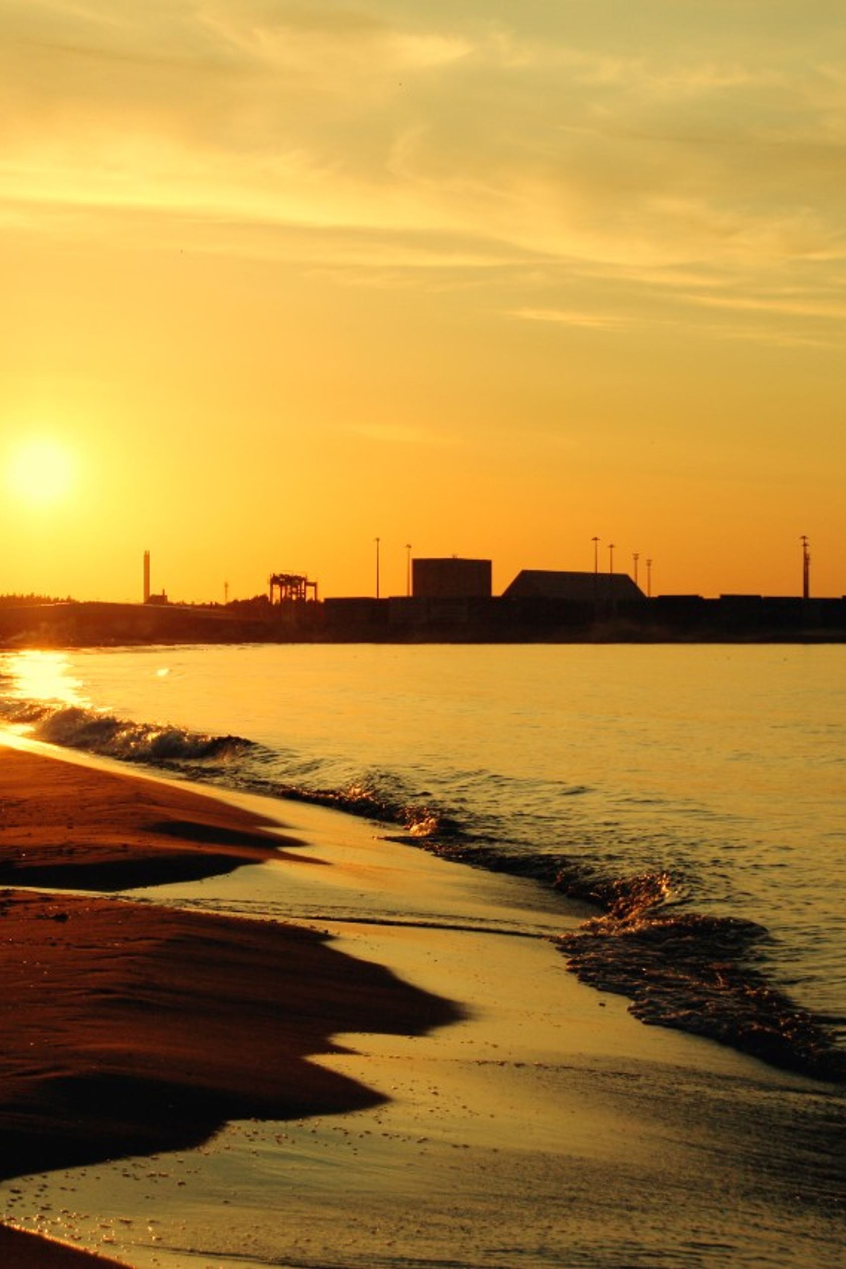 Sonnenuntergang über einem ruhigen Strand mit sanften Wellen, im Hintergrund Industriekräne und Gebäude, die eine harmonische Verbindung von Natur und urbanem Hafen zeigen.