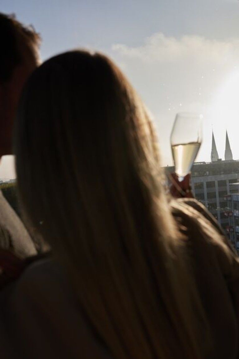 Ein Paar genießt bei Sonnenuntergang einen Drink auf einer Terrasse mit Blick auf eine Stadt und zwei Kirchturmspitzen im Hintergrund.