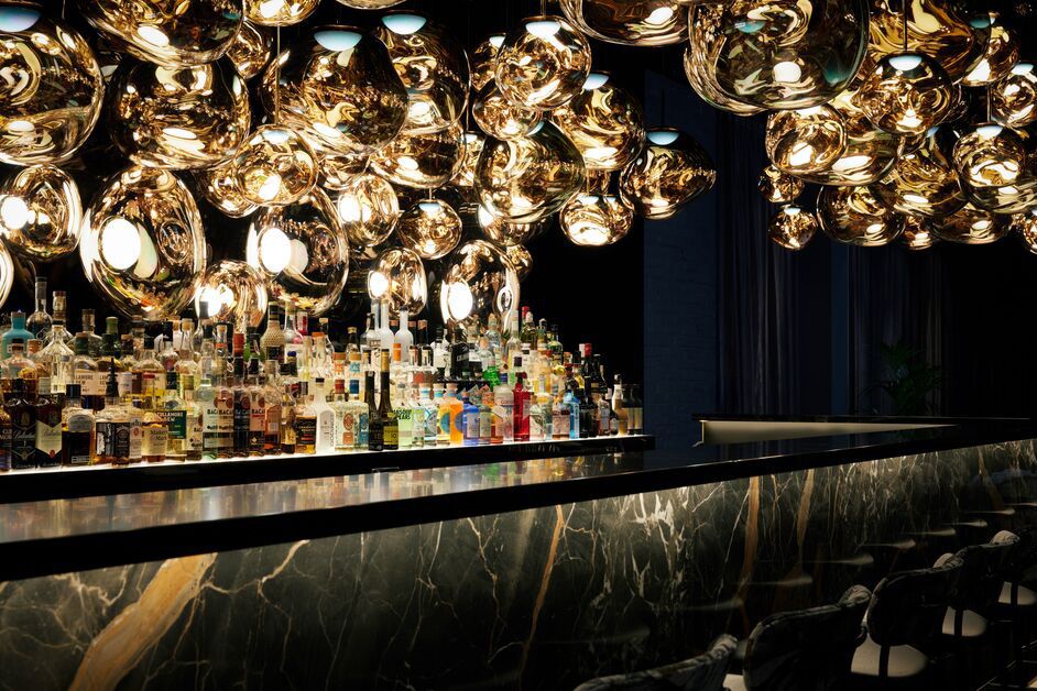Elegante Hotelbar mit gedämpfter Beleuchtung, glänzenden goldfarbenen Deckenlichtern und einer gut bestückten Spirituosenauswahl hinter einer marmornen Bar. Gemütliche Barhocker laden zum Verweilen ein.