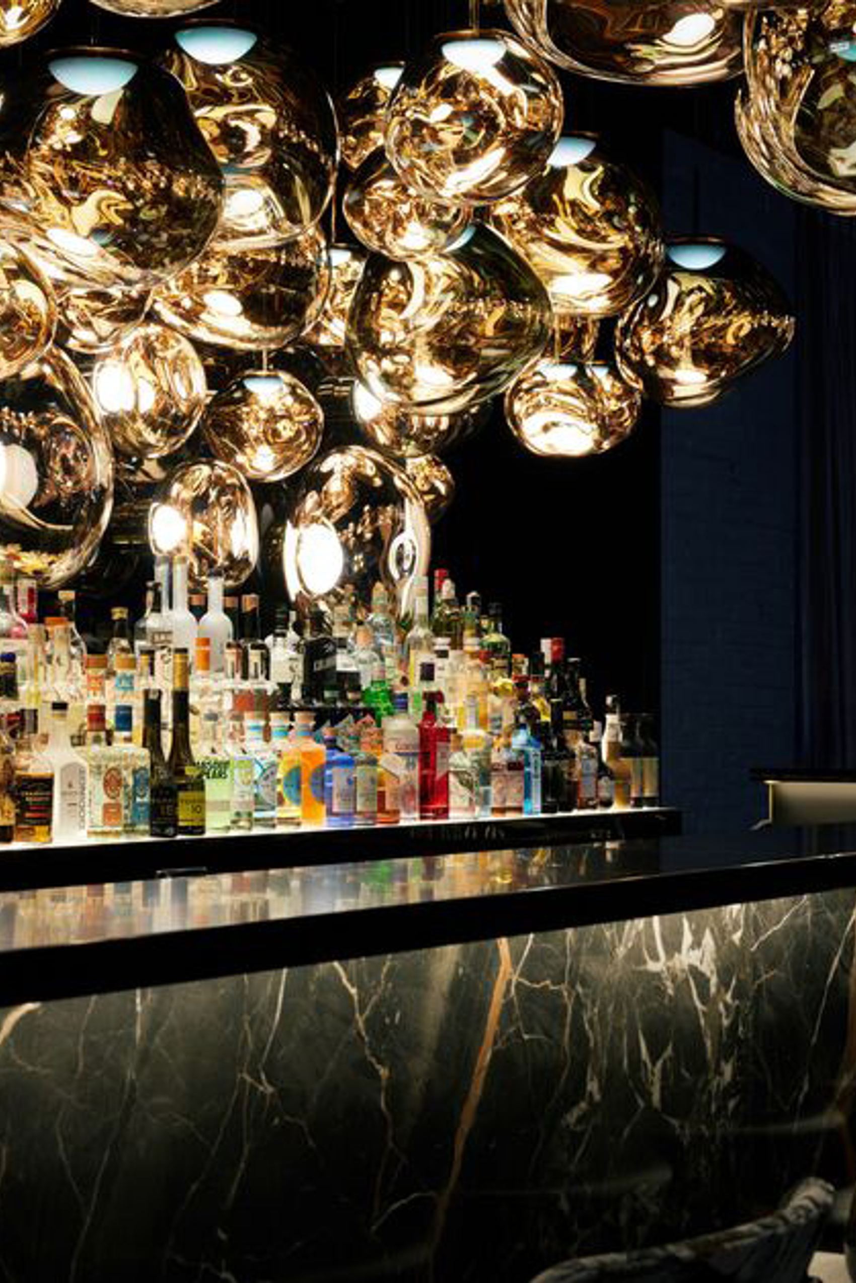 Elegante Hotelbar mit gedämpfter Beleuchtung, glänzenden goldfarbenen Deckenlichtern und einer gut bestückten Spirituosenauswahl hinter einer marmornen Bar. Gemütliche Barhocker laden zum Verweilen ein.