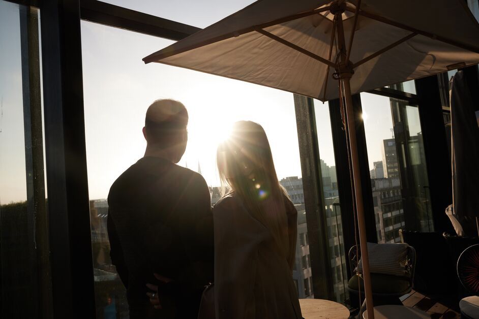 Zwei Personen genießen den Sonnenuntergang gemeinsam auf einer Hotelterrasse mit großem Sonnenschirm und Blick über die Stadt.
