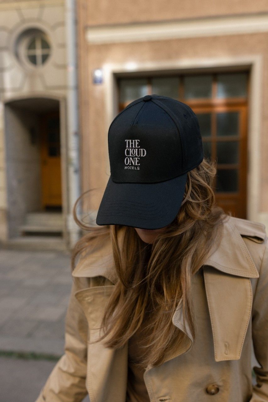 Frau mit langer, wehender Haarpracht trägt eine schwarze Baseballkappe mit der Aufschrift „THE CLOUD ONE HOTEL“ und einen beigen Trenchcoat vor einem urbanen Gebäude.