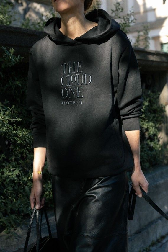 Person trägt schwarzen Kapuzenpullover mit Aufdruck „THE CLOUD ONE HOTELS“ und schwarze Lederhose, geht draußen spazierend, hält Taschen in beiden Händen.