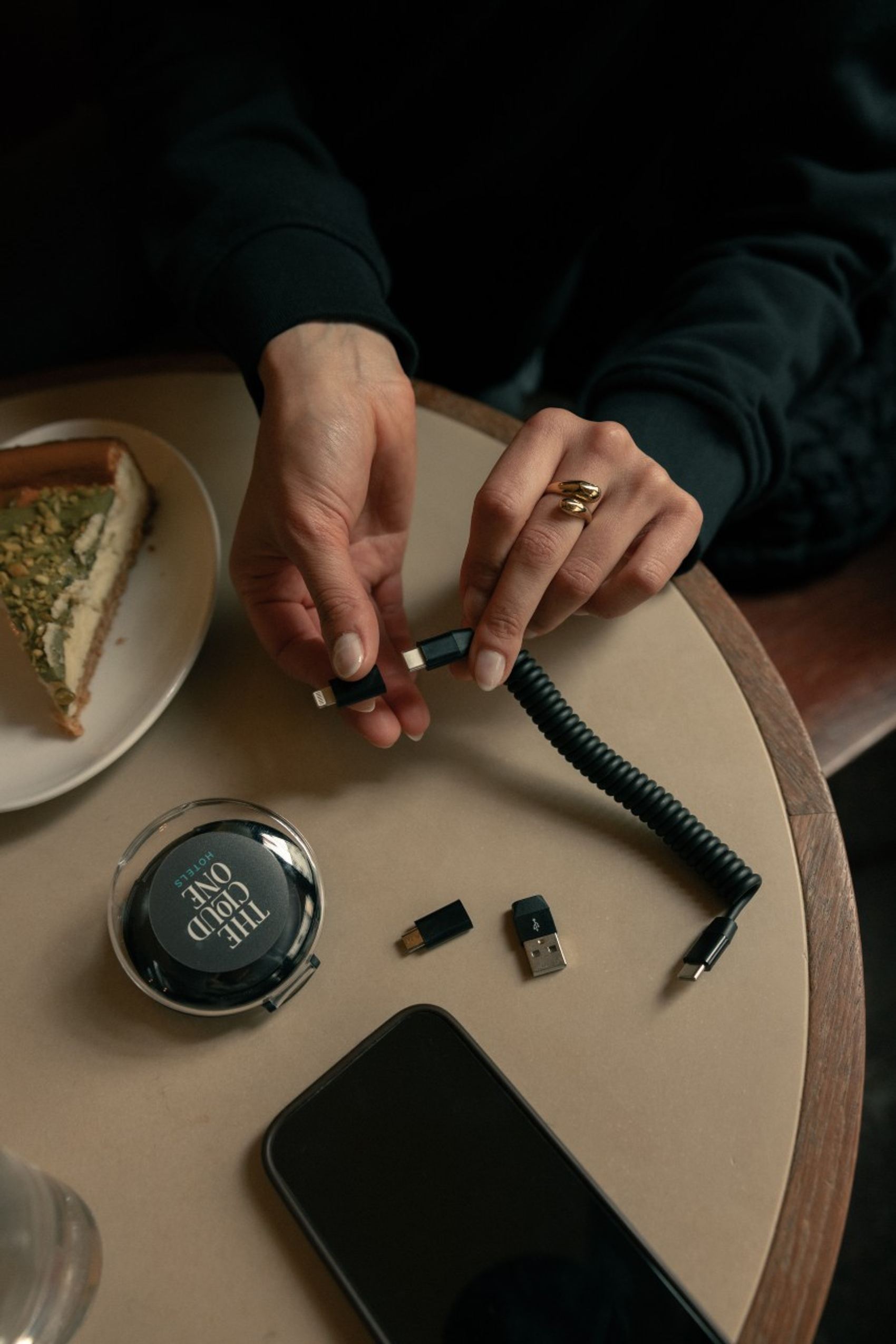Hände verbinden ein kurzes, spiralgestricktes Ladekabel. Auf dem Tisch liegen ein Smartphone, ein USB-Adapter, eine runde Dose mit der Aufschrift "THE WILD ONE" und ein Stück Kuchen auf einem Teller.