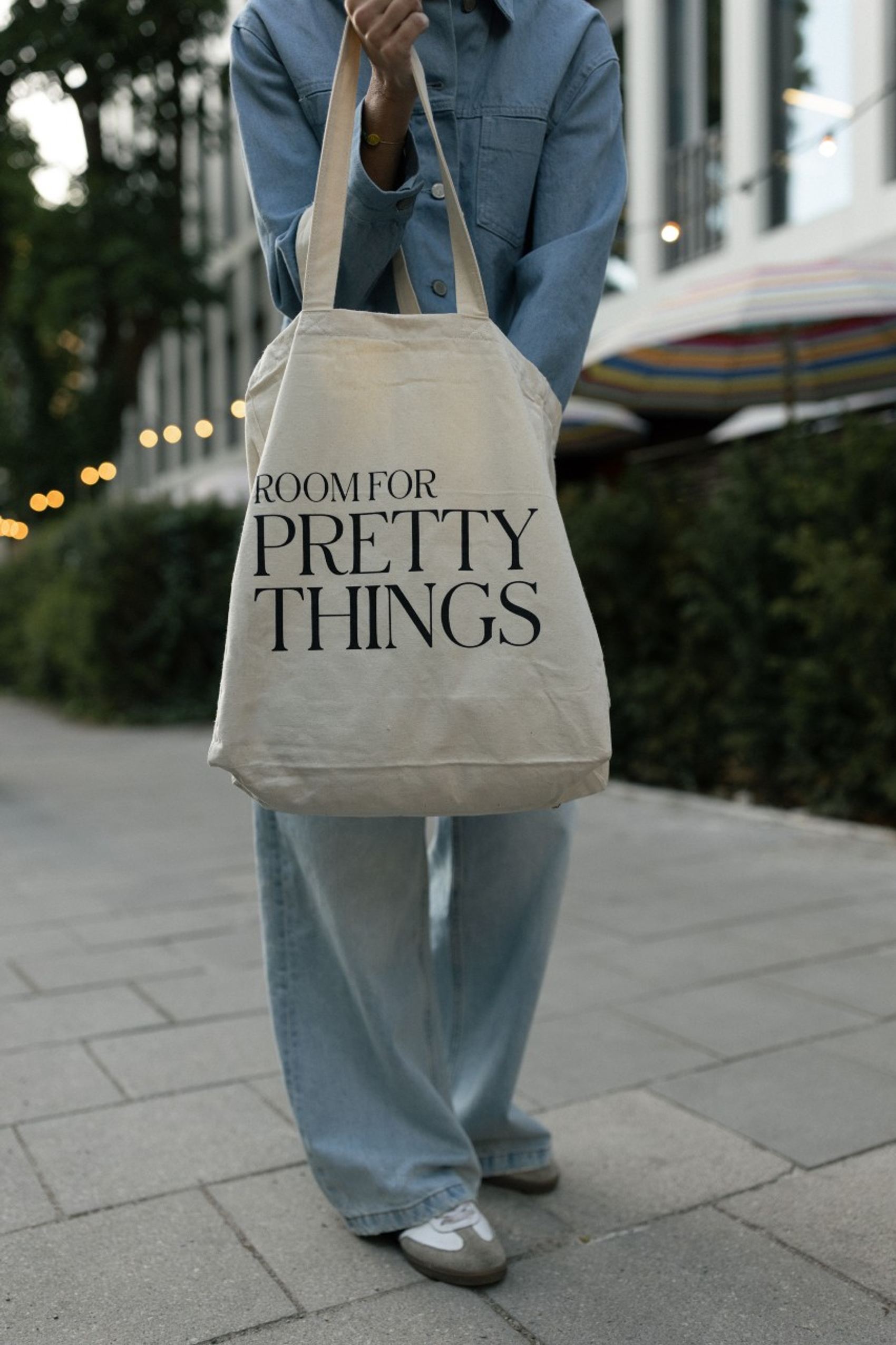 Person hält eine Baumwolltasche mit dem Aufdruck „ROOM FOR PRETTY THINGS“, bekleidet mit blauer Jeans und Hemd, stehend auf einem Gehweg in urbaner Umgebung.