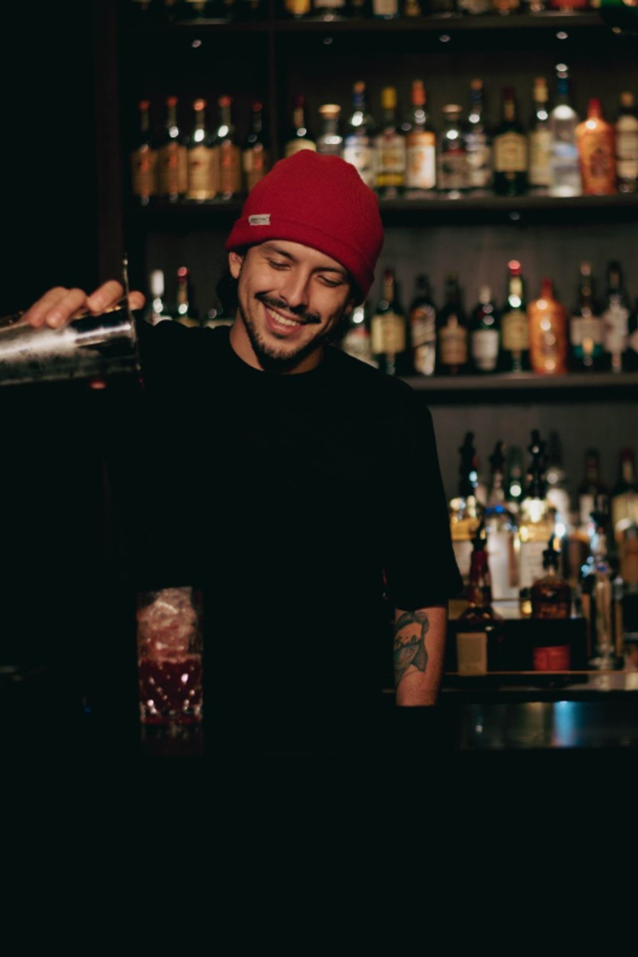 Barkeeper mit roter Mütze mischt lächelnd einen Cocktail vor einer gut bestückten Bar, die in warmem Licht erstrahlt.
