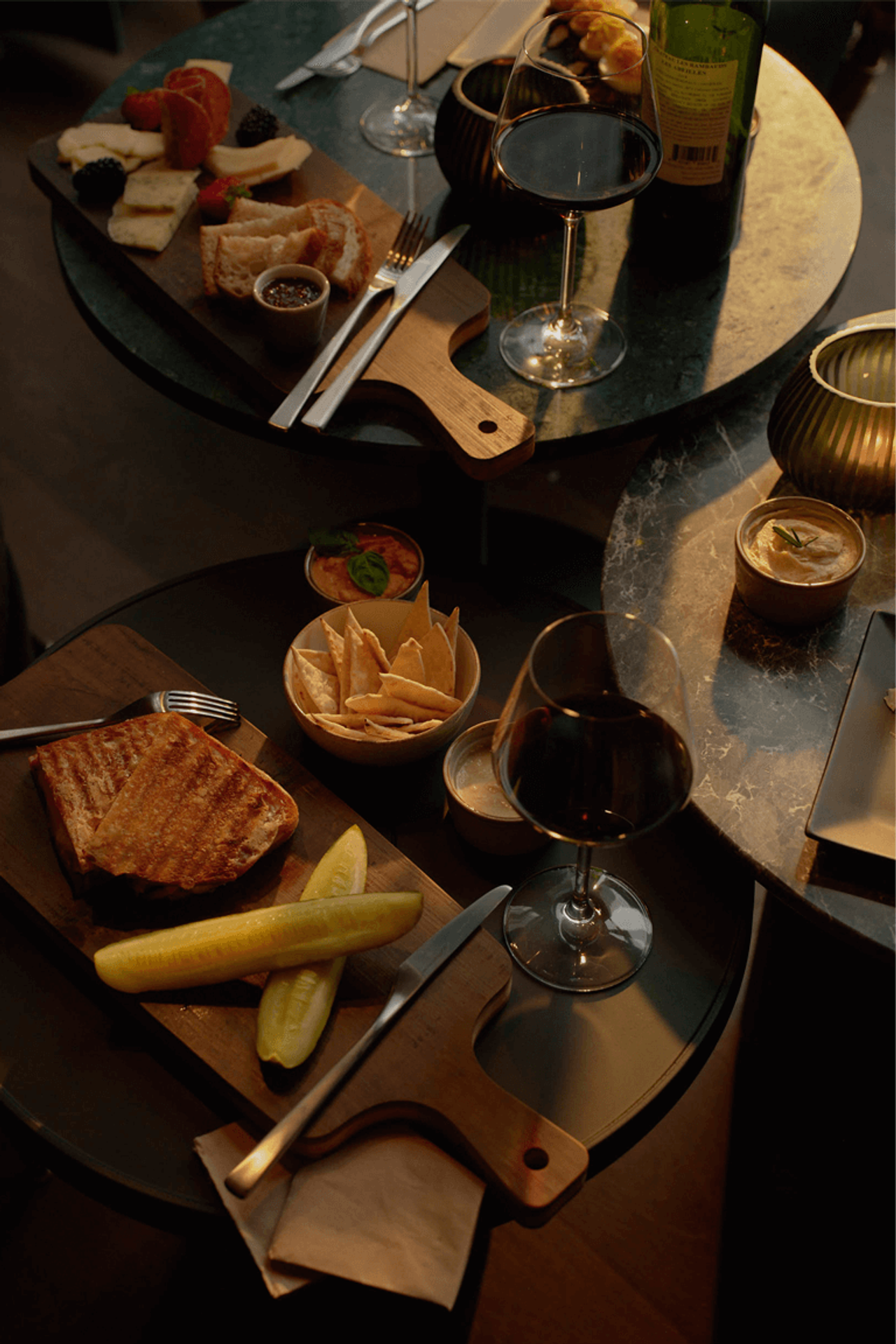 Zwei Holzbretter mit Snacks wie Käse, Brot, Gurken und Chips, dazu zwei Gläser Rotwein auf einem dunklen Tisch