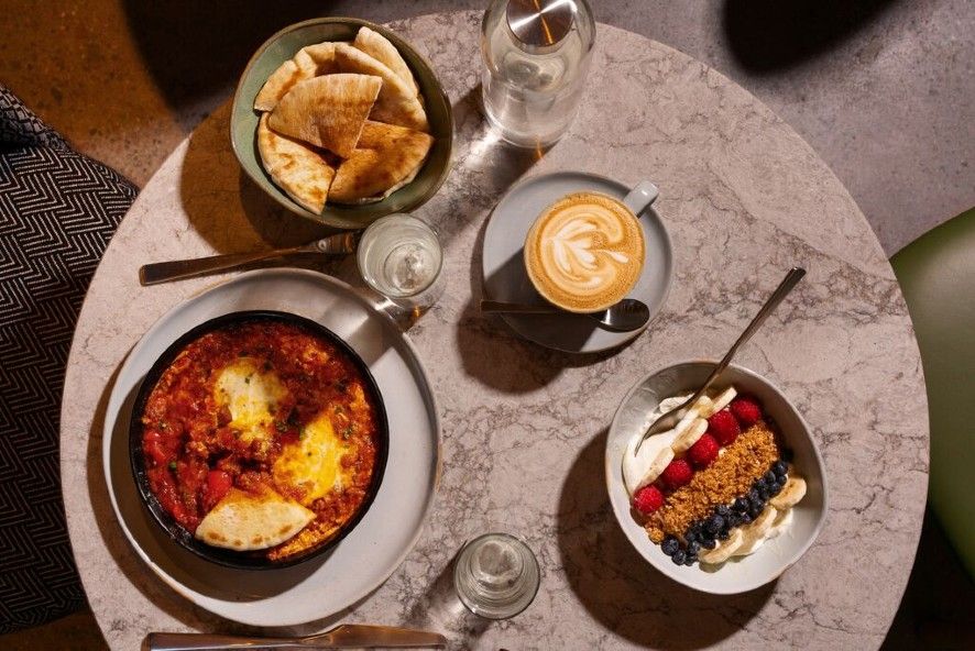 Ein gedeckter Tisch mit Shakshuka in einer Pfanne, einem Becher mit Cappuccino, einem Schälchen mit Pita-Brot und einer Schüssel mit Joghurt, Beeren und Granola, perfekt für ein ausgewogenes, genussvolles Frühstück.