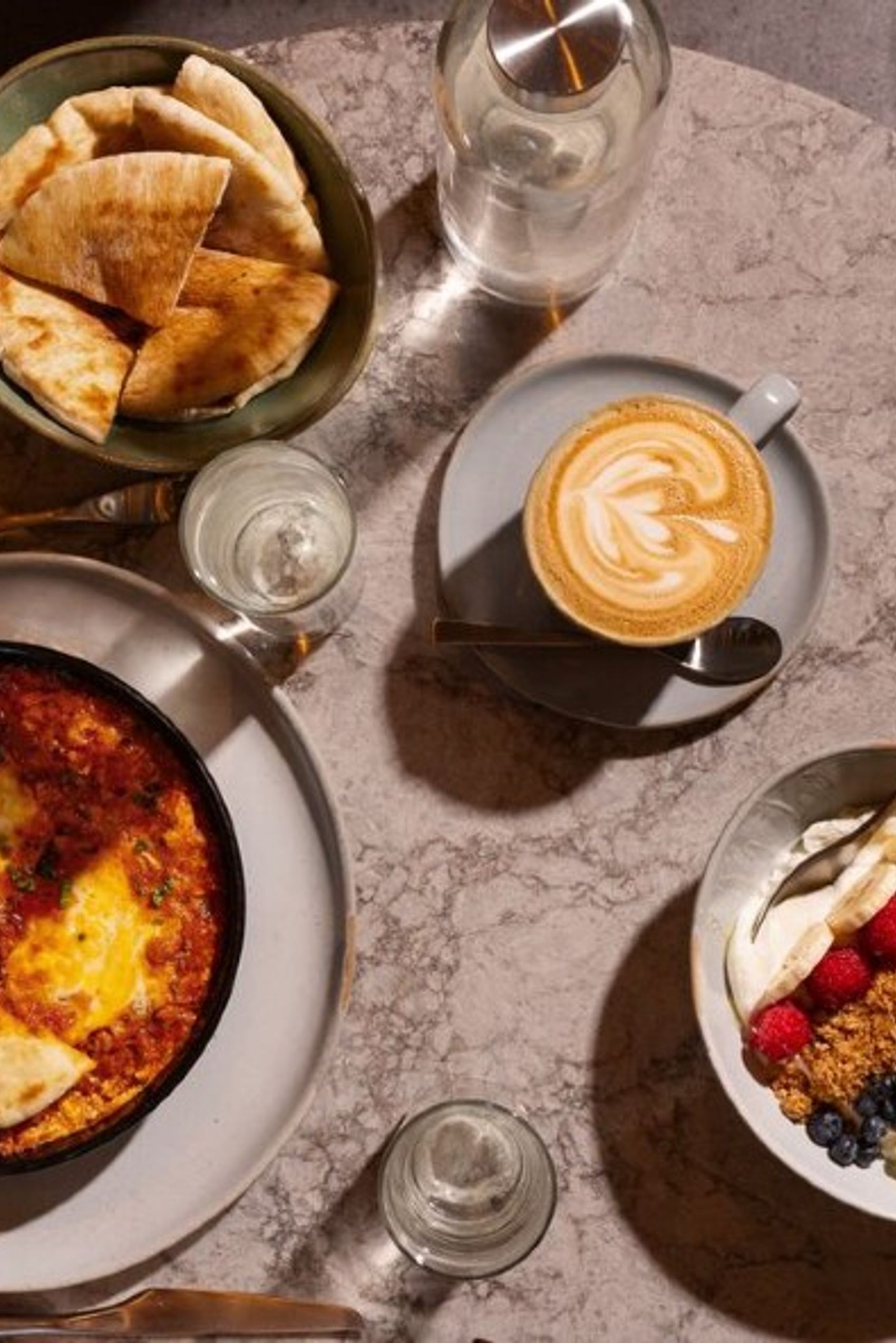 Ein gedeckter Tisch mit Shakshuka in einer Pfanne, einem Becher mit Cappuccino, einem Schälchen mit Pita-Brot und einer Schüssel mit Joghurt, Beeren und Granola, perfekt für ein ausgewogenes, genussvolles Frühstück.