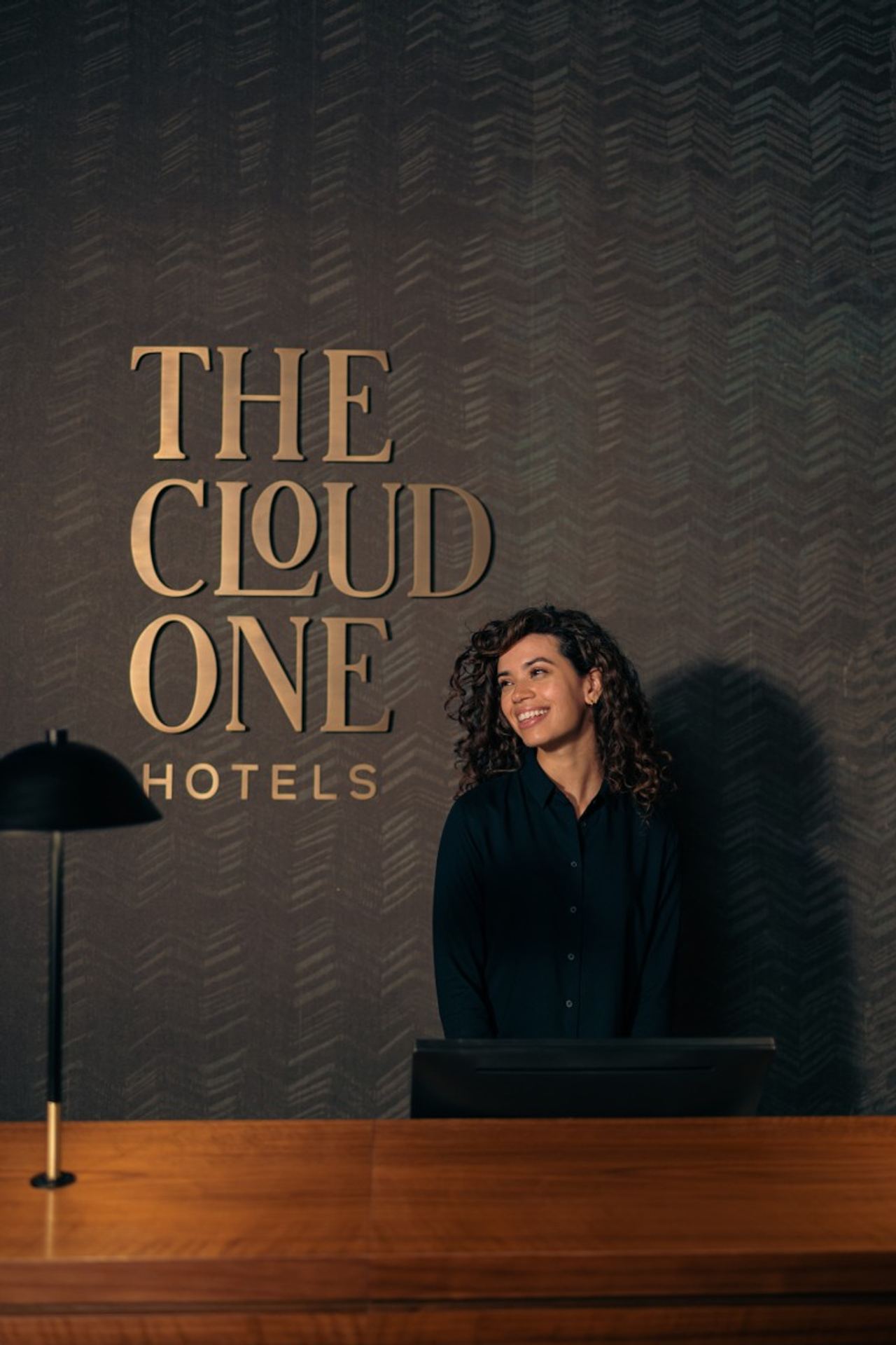 Rezeptionistin lächelt freundlich hinter der Empfangstheke eines stilvollen Hotels mit dunkel gestaltetem Hintergrund und großem Schriftzug „THE CLOUD ONE HOTELS“.