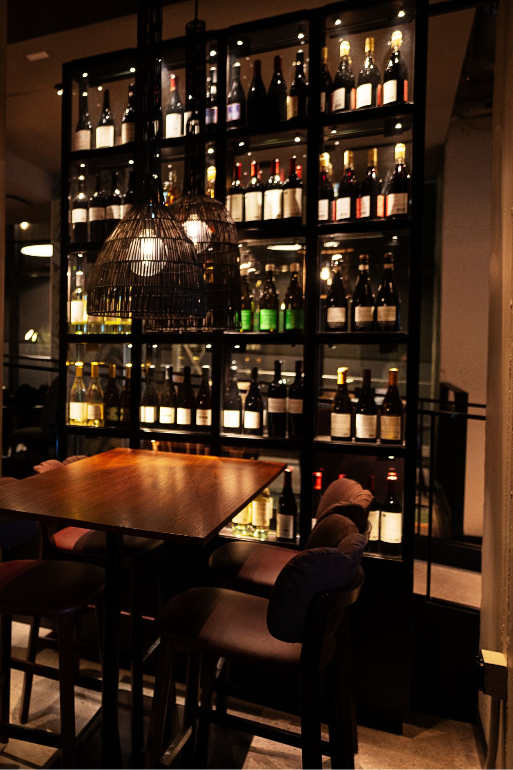 The Cloud One Deli gemütliche Bar-Ecke mit Holztisch und bequemen Stühlen, umgeben von einem beleuchteten Regal voller Weinflaschen, das eine elegante und einladende Atmosphäre für genussvolle Momente schafft.