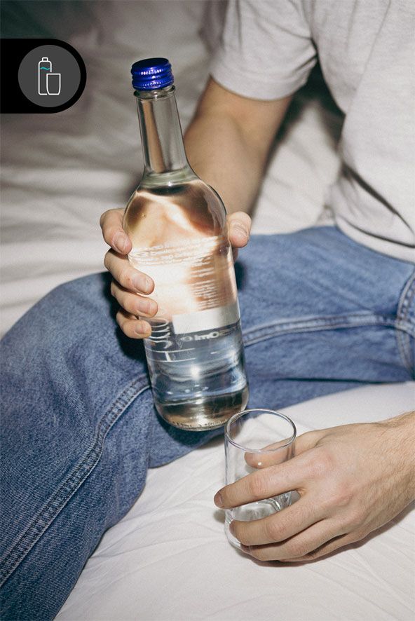 Person sitzt auf Bett und hält eine Glasflasche mit Wasser in einer Hand, in der anderen ein leeres Glas zum Einschenken.
