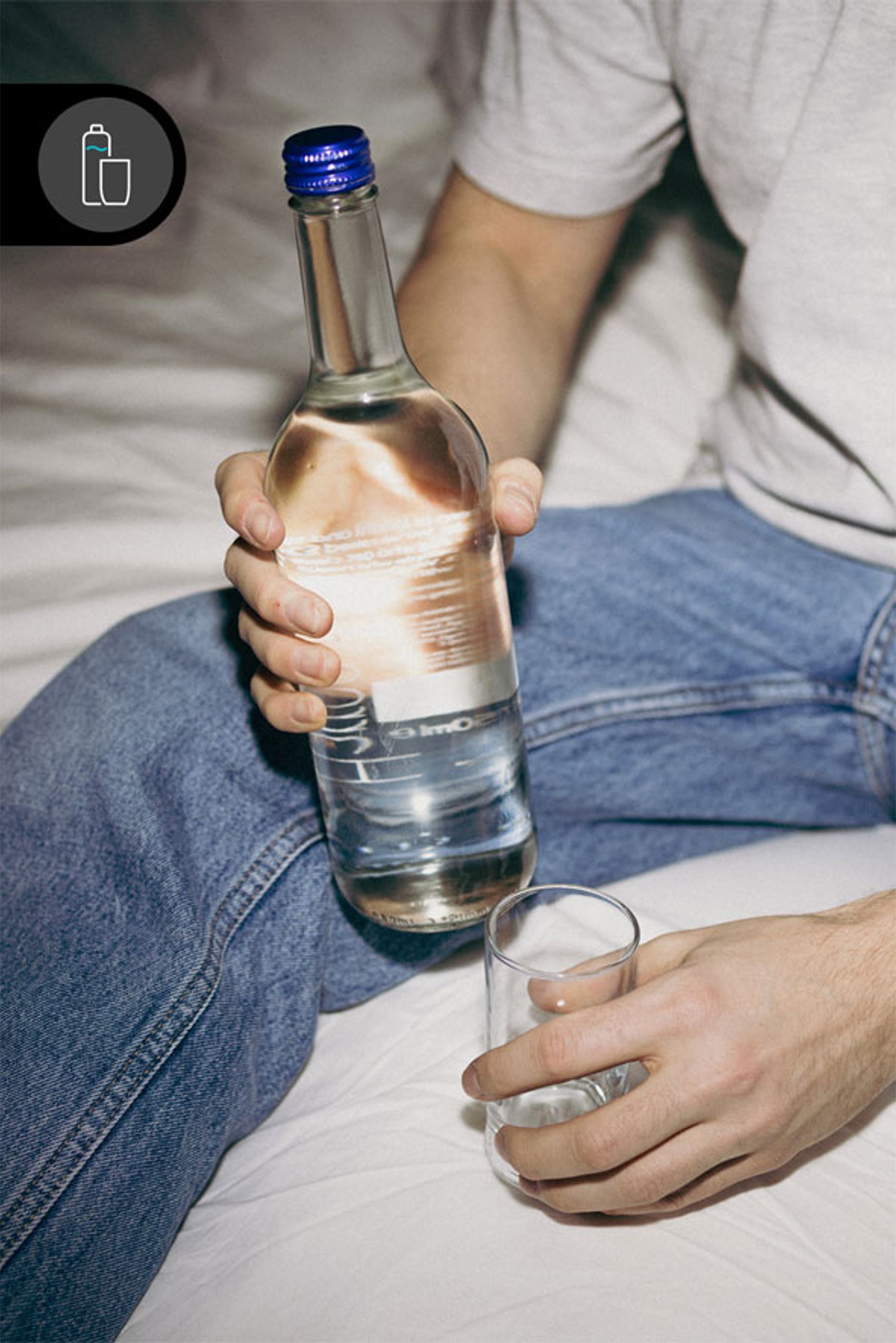 Person sitzt auf Bett und hält eine Glasflasche mit Wasser in einer Hand, in der anderen ein leeres Glas zum Einschenken.