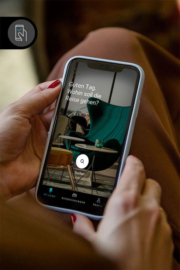 Person hält ein Smartphone mit einer Reisebuchungs-App, die auf dem Bildschirm die Frage "Guten Tag. Wohin soll die Reise gehen?" anzeigt. Im Hintergrund ist ein gemütlicher Raum mit einem Stuhl und Tisch zu sehen. Person hält ein Smartphone mit einer Reisebuchungs-App, die auf dem Bildschirm die Frage "Guten Tag. Wohin soll die Reise gehen?" anzeigt. Im Hintergrund ist ein gemütlicher Raum mit einem Stuhl und Tisch zu sehen.