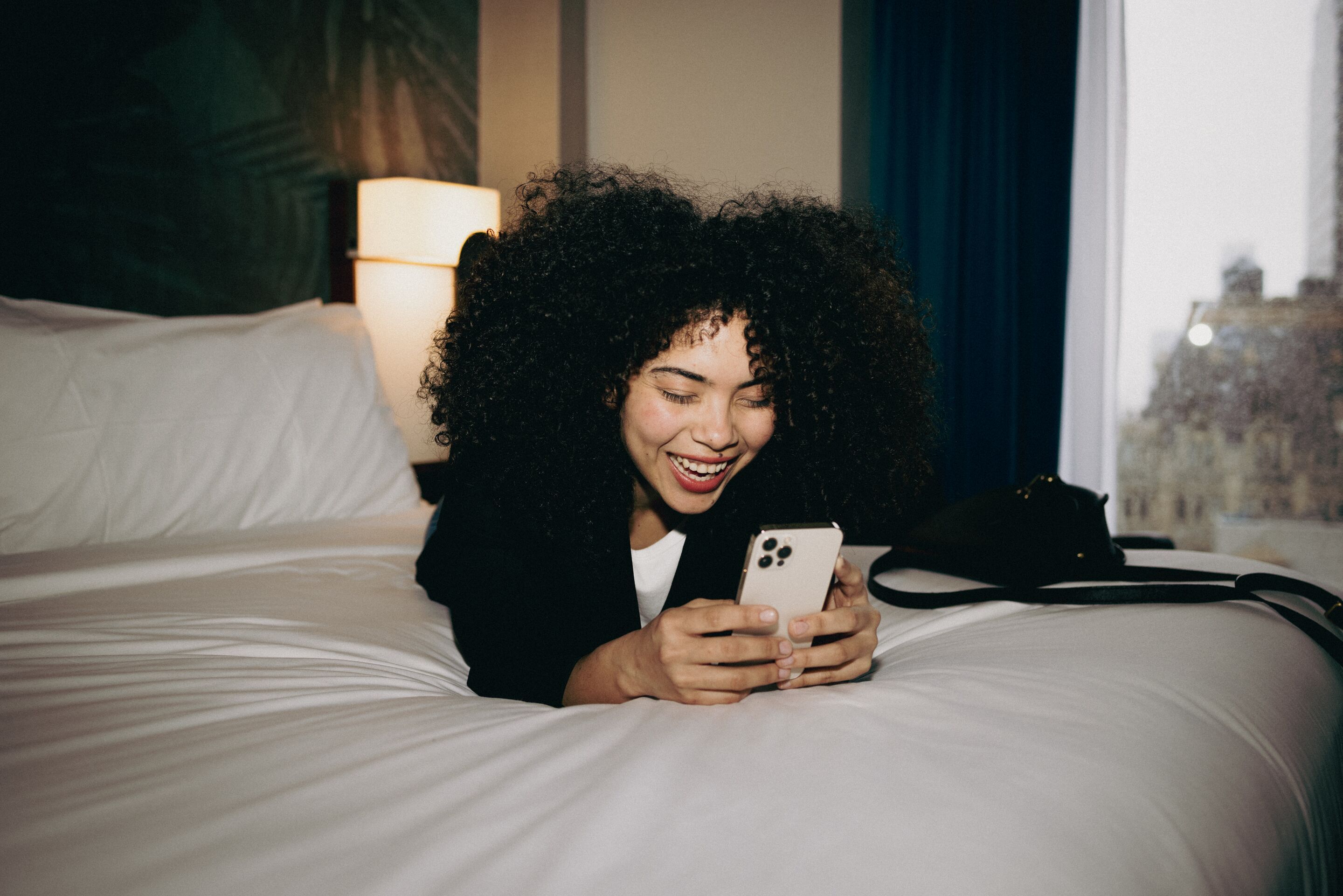 Frau liegt entspannt auf einem Hotelbett und lächelt beim Blick auf ihr Smartphone, gemütliche Atmosphäre im Zimmer mit warmem Licht.