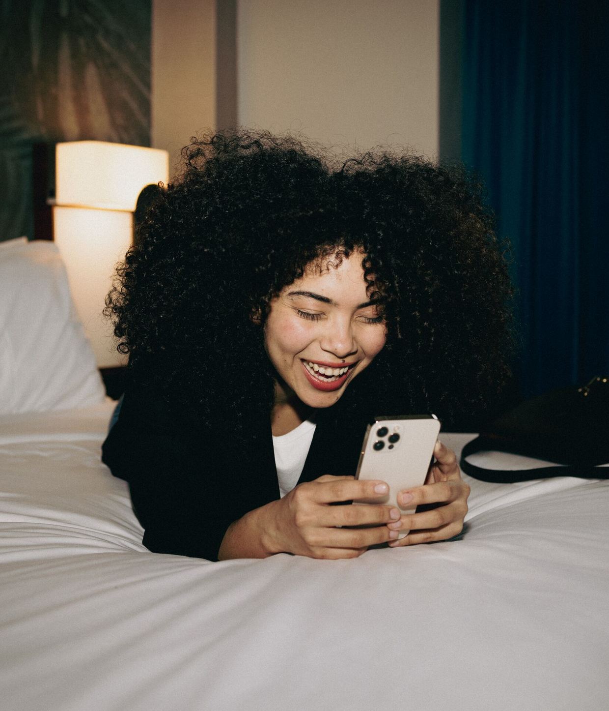 Frau liegt entspannt auf einem Hotelbett und lächelt beim Blick auf ihr Smartphone, gemütliche Atmosphäre im Zimmer mit warmem Licht.