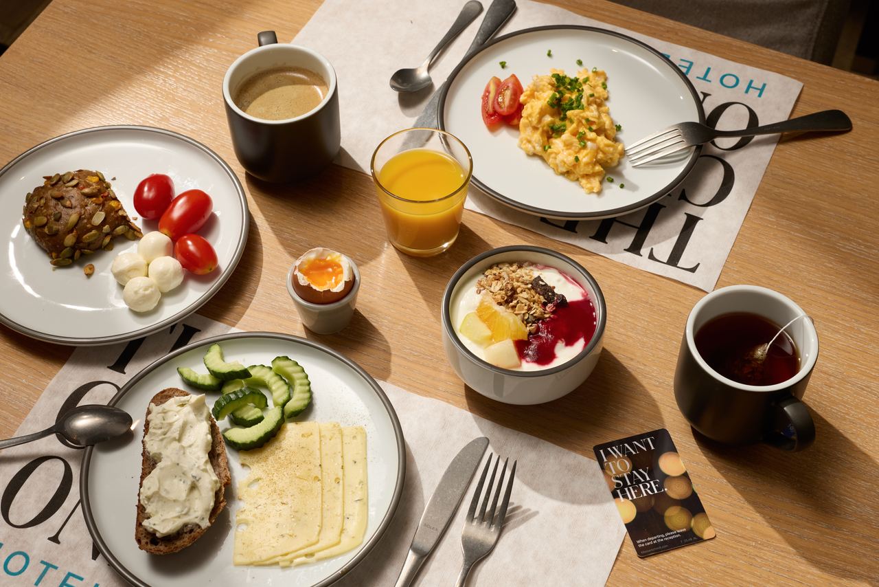 Frühstück mit Rührei, Joghurt mit Müsli und Beeren, Käse, Frischkäsebrot, Tomaten, Gurkenscheiben, gekochtem Ei, Orangensaft, Kaffee und Tee auf hellem Holztisch.