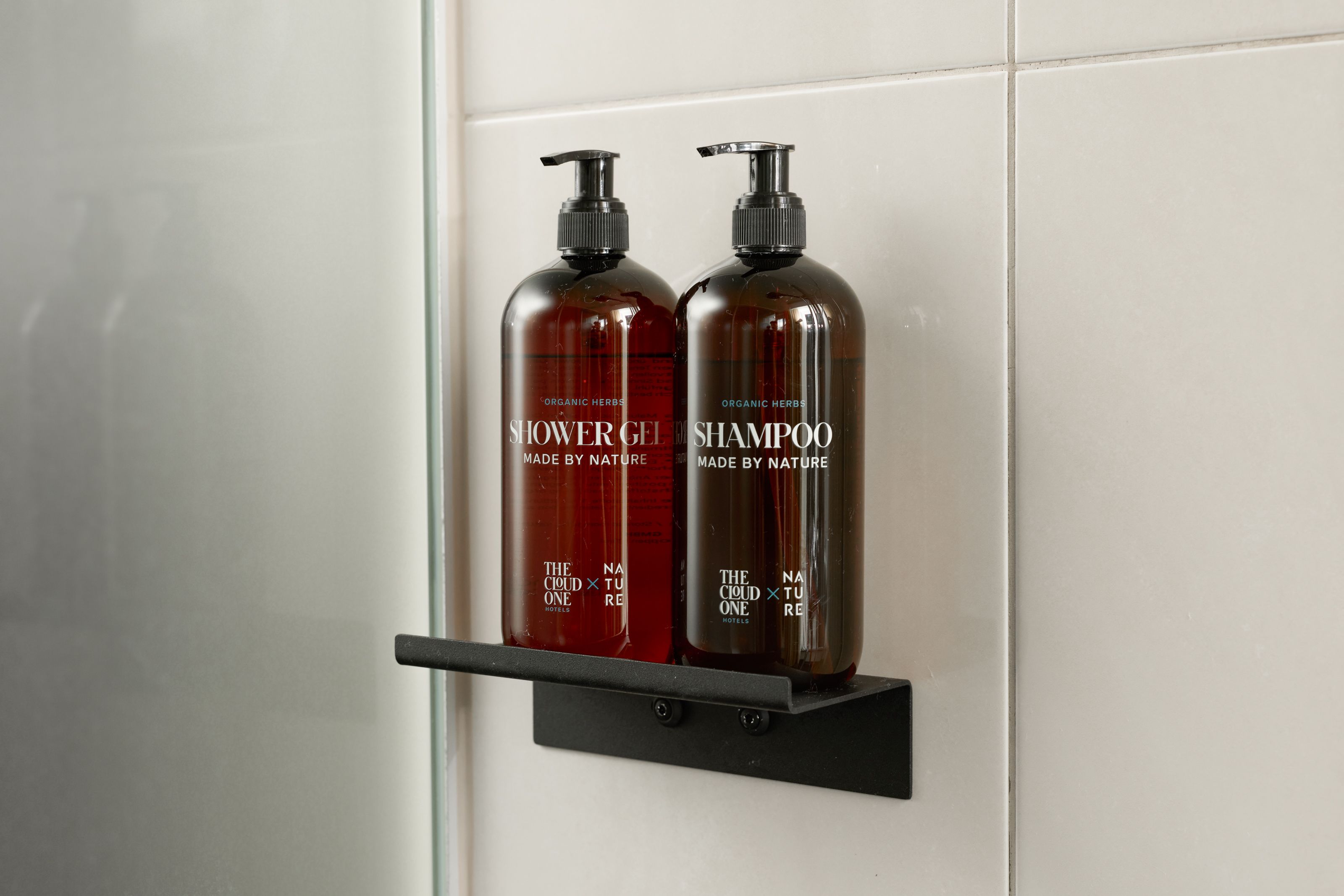 Zwei braune Flaschen mit Shampoo und Duschgel, jeweils mit Pumpspender, stehen auf einem schwarzen Regal in einer modernen Duschkabine vor weißen Wandfliesen.