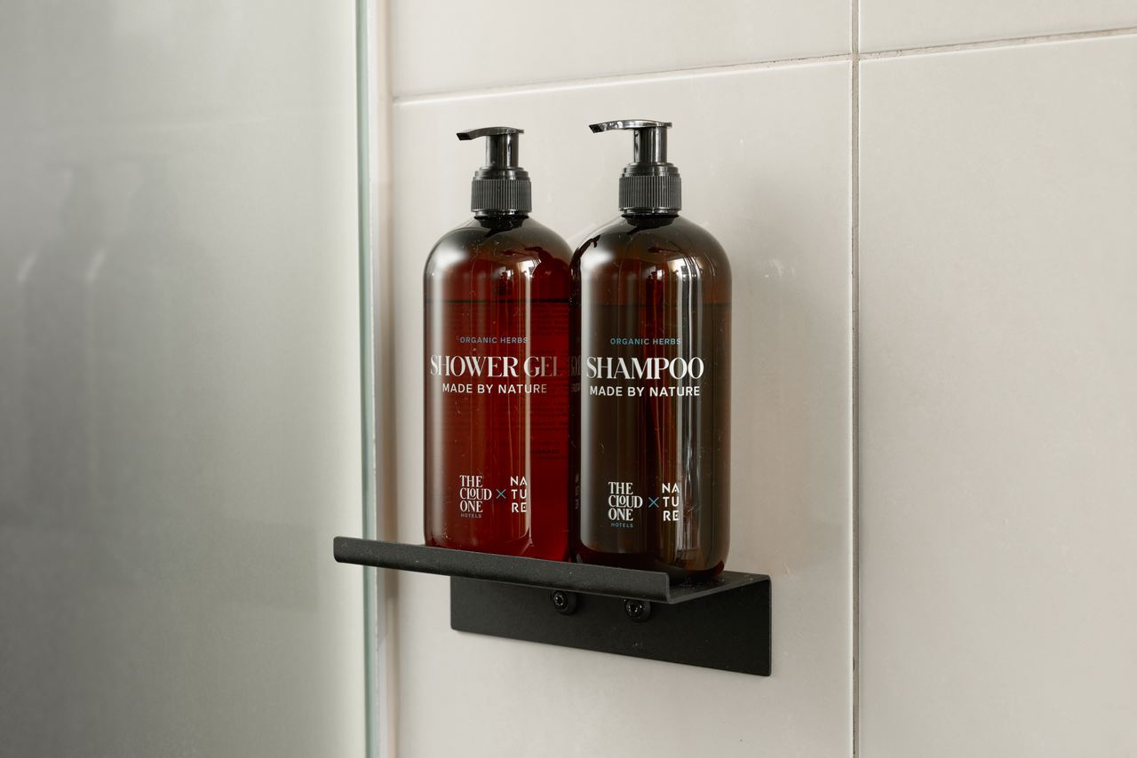Zwei braune Flaschen mit Shampoo und Duschgel, jeweils mit Pumpspender, stehen auf einem schwarzen Regal in einer modernen Duschkabine vor weißen Wandfliesen.