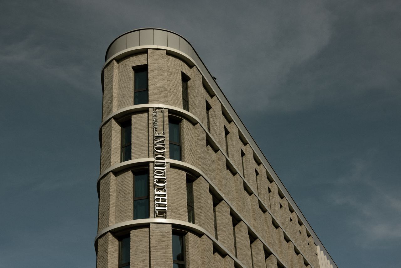 Ansicht eines modernen, hellen Hotelgebäudes mit abgerundeter Fassade und vertikalem Schriftzug „THE CLOUD ON“. Klare Linien und große Fenster verleihen dem Haus ein elegantes, zeitgemäßes Erscheinungsbild vor bewölktem Himmel.