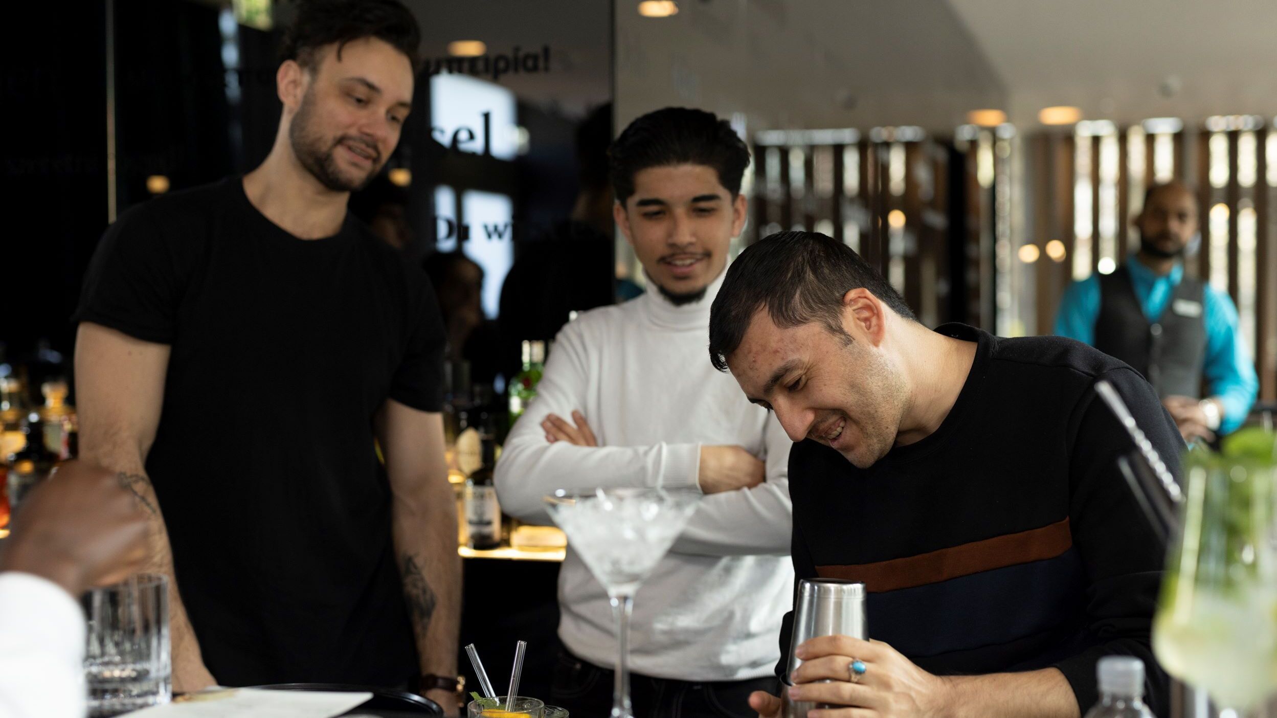 Drei Männer beobachten interessiert, wie ein Barkeeper einen Cocktail zubereitet, und genießen die entspannte Atmosphäre an der Bar.