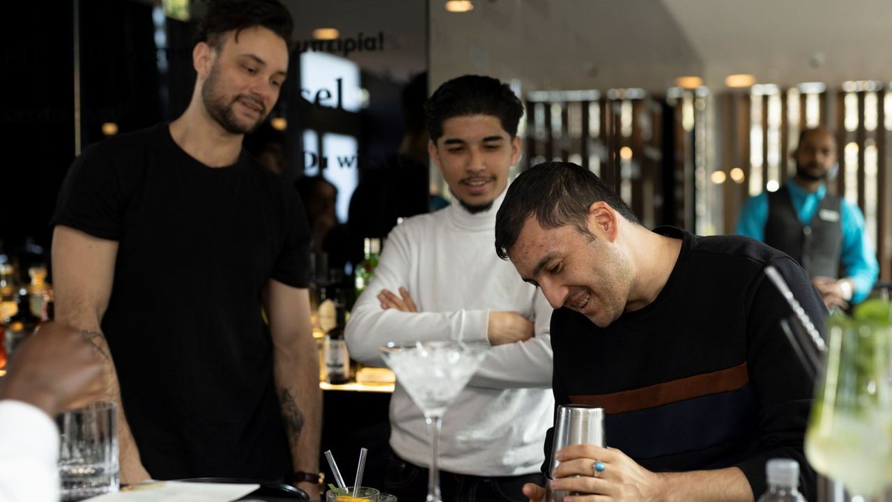 Drei Männer beobachten interessiert, wie ein Barkeeper einen Cocktail zubereitet, und genießen die entspannte Atmosphäre an der Bar.