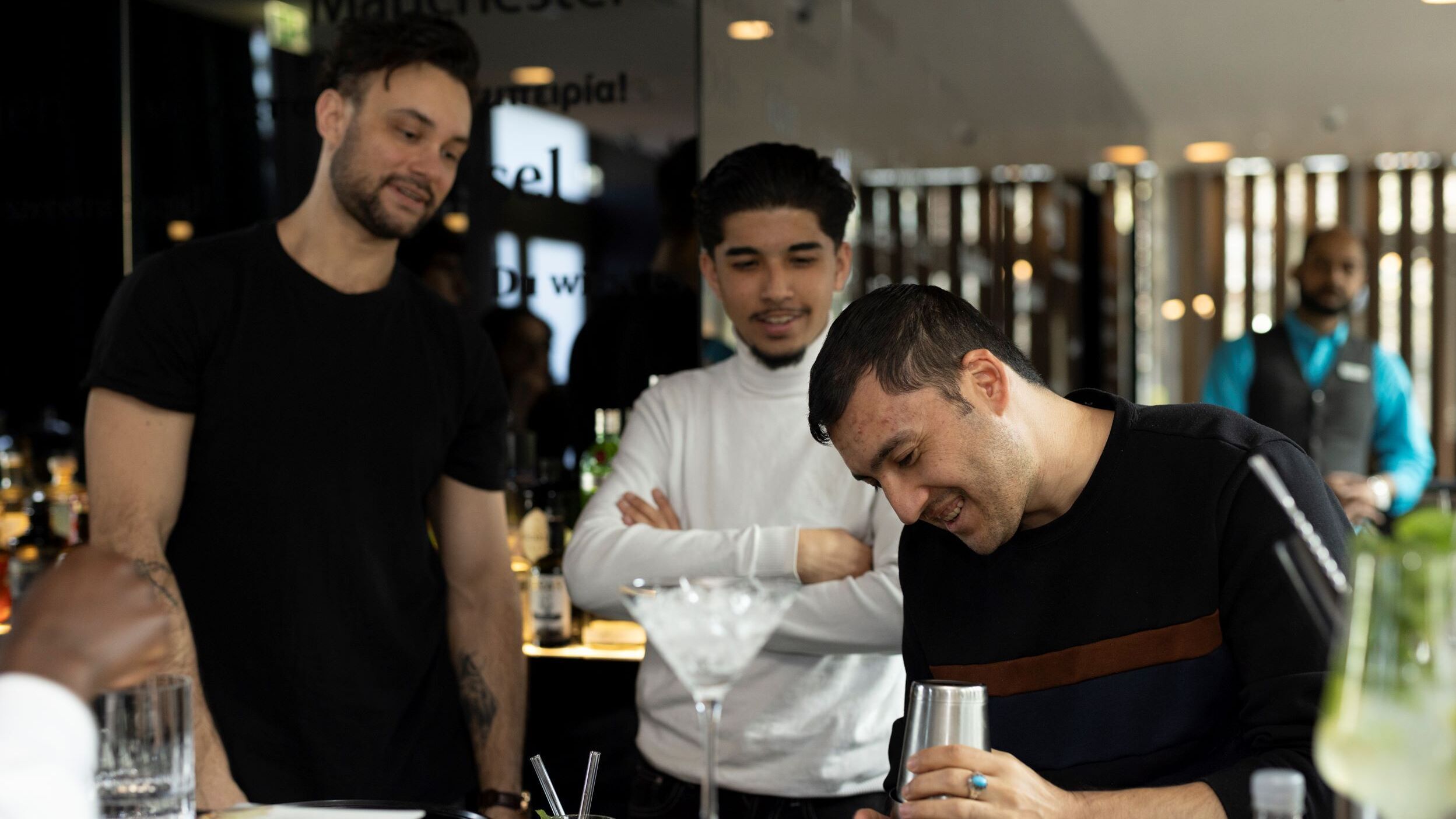 Drei Männer beobachten interessiert, wie ein Barkeeper einen Cocktail zubereitet, und genießen die entspannte Atmosphäre an der Bar.