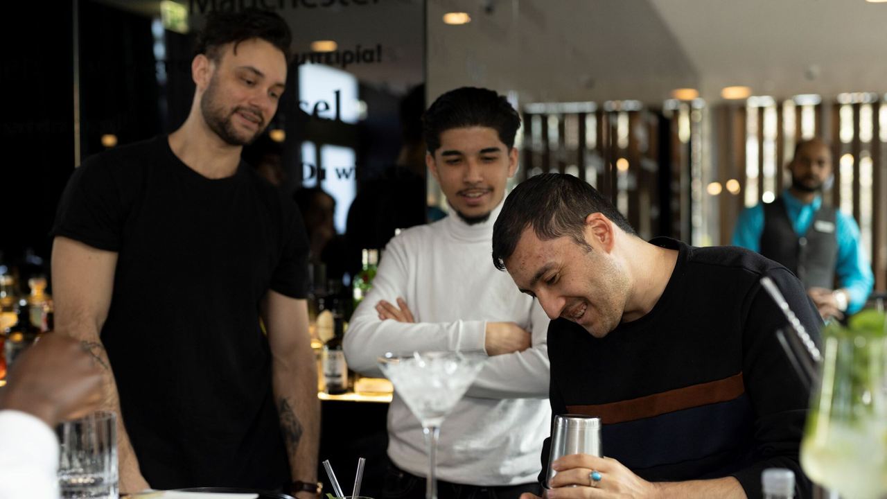 Drei Männer beobachten interessiert, wie ein Barkeeper einen Cocktail zubereitet, und genießen die entspannte Atmosphäre an der Bar. Drei Männer beobachten interessiert, wie ein Barkeeper einen Cocktail zubereitet, und genießen die entspannte Atmosphäre an der Bar.