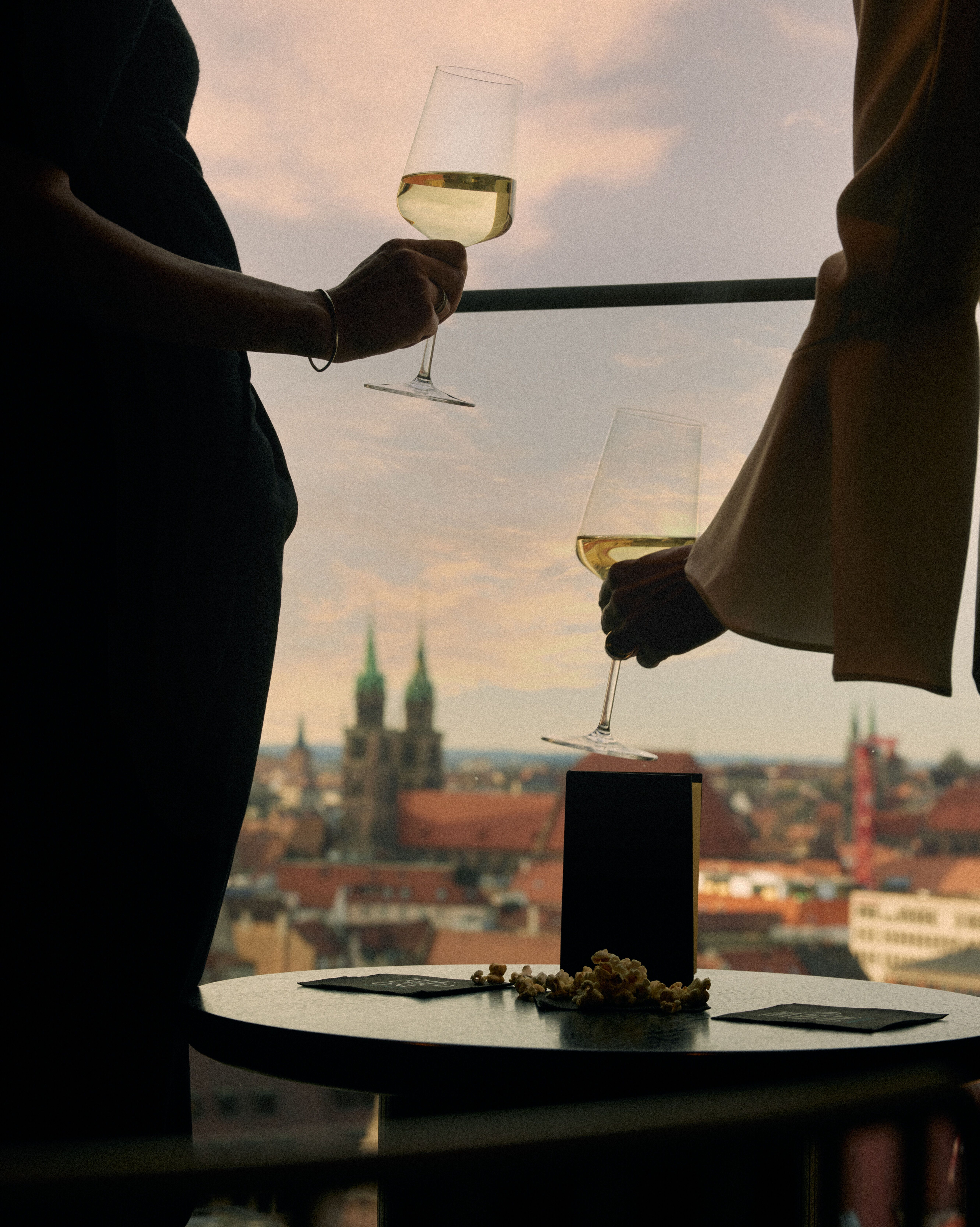Zwei Personen genießen bei Sonnenuntergang Weißwein auf einem Balkon mit Blick auf eine historische Stadtlandschaft.