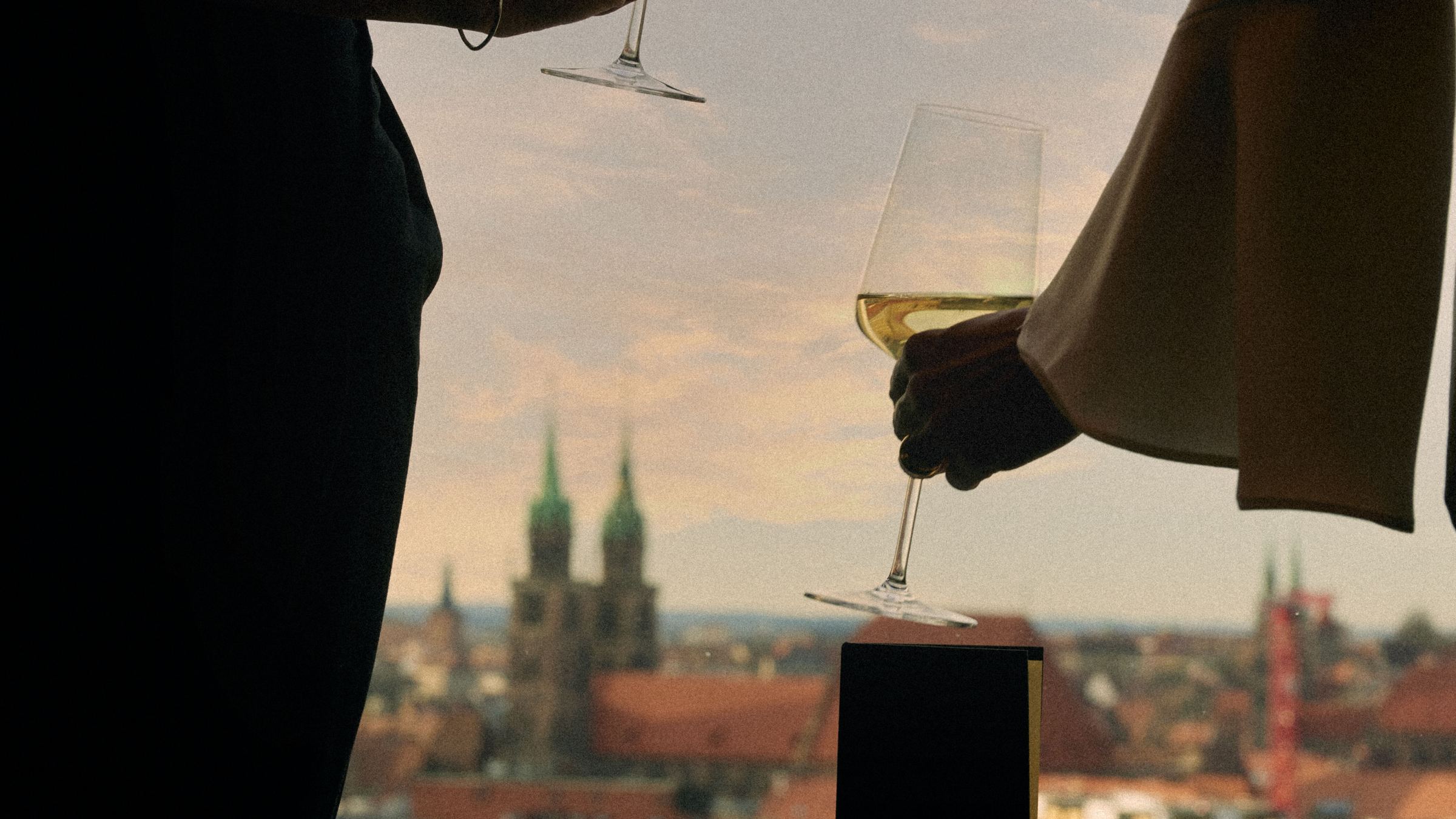 Zwei Personen genießen bei Sonnenuntergang Weißwein auf einem Balkon mit Blick auf eine historische Stadtlandschaft. Zwei Personen genießen bei Sonnenuntergang Weißwein auf einem Balkon mit Blick auf eine historische Stadtlandschaft.