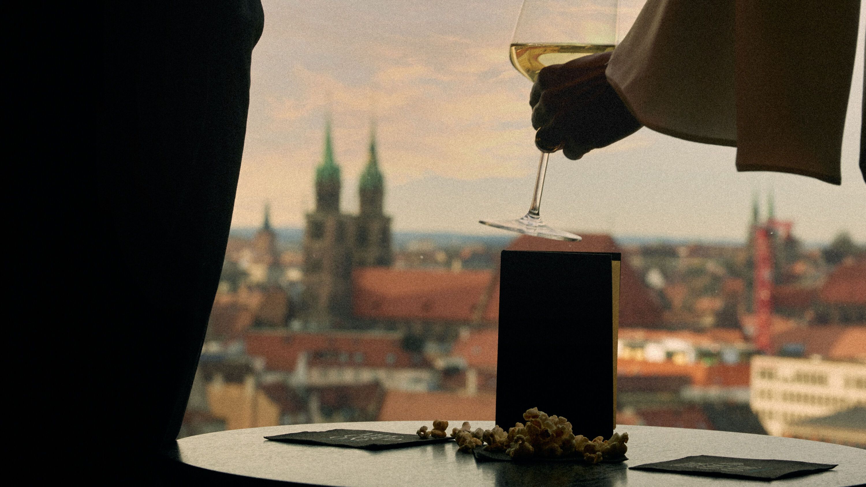 Zwei Personen genießen bei Sonnenuntergang Weißwein auf einem Balkon mit Blick auf eine historische Stadtlandschaft.