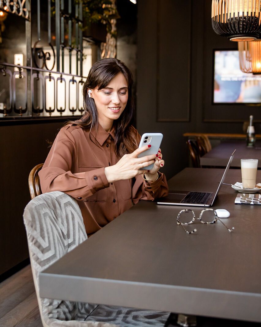 Frau sitzt entspannt an einem großen Tisch in modernem Café, nutzt Smartphone, neben ihr Laptop und Getränke. Gemütliche Atmosphäre, geeignet für Arbeit und Freizeit.