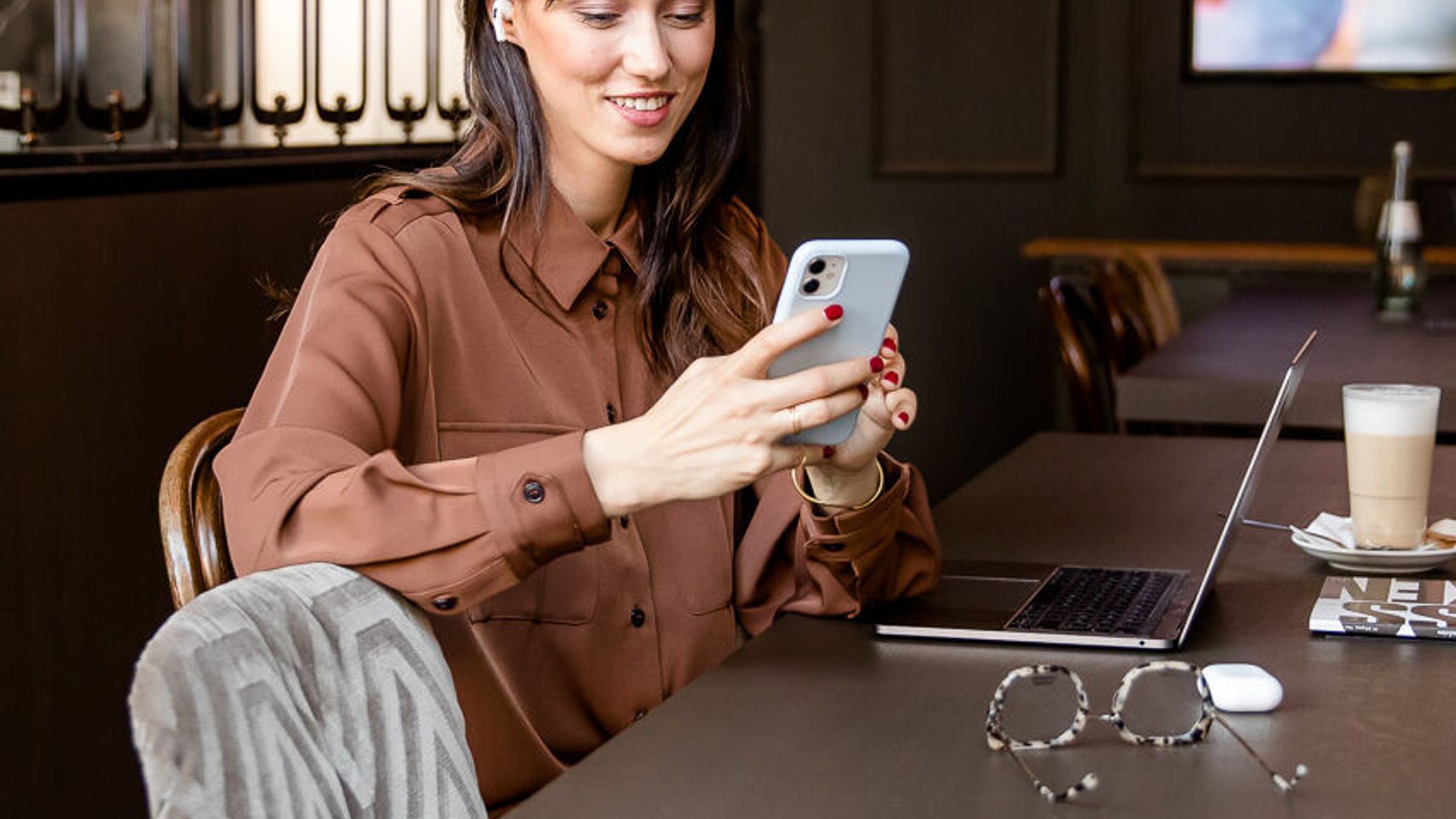 Frau sitzt entspannt an einem großen Tisch in modernem Café, nutzt Smartphone, neben ihr Laptop und Getränke. Gemütliche Atmosphäre, geeignet für Arbeit und Freizeit. Frau sitzt entspannt an einem großen Tisch in modernem Café, nutzt Smartphone, neben ihr Laptop und Getränke. Gemütliche Atmosphäre, geeignet für Arbeit und Freizeit.