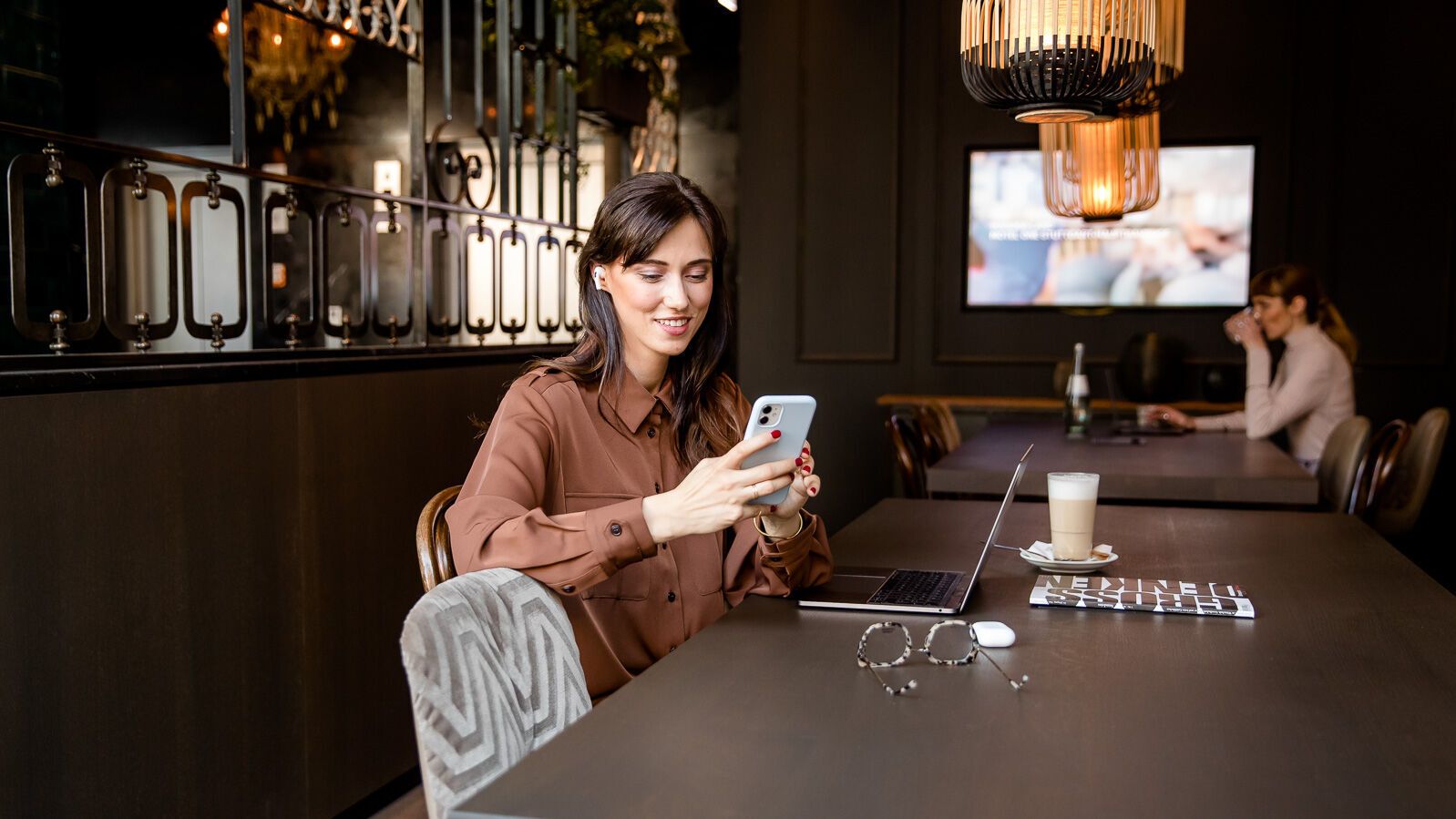 Frau sitzt entspannt an einem großen Tisch in modernem Café, nutzt Smartphone, neben ihr Laptop und Getränke. Gemütliche Atmosphäre, geeignet für Arbeit und Freizeit.