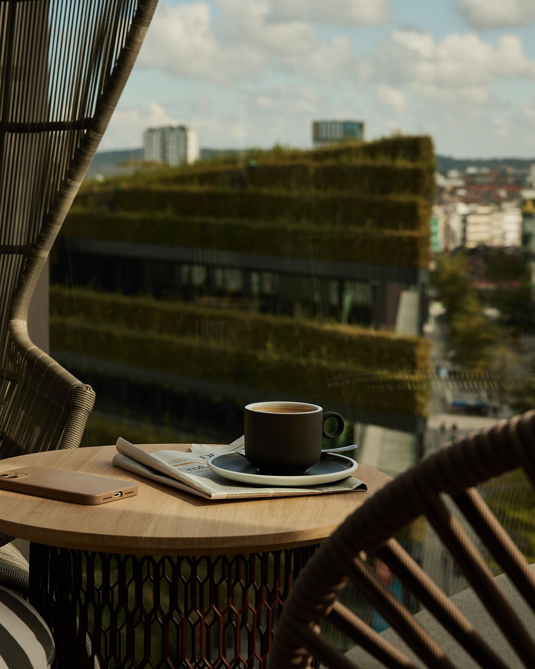 Balkon mit bequemen Stühlen und kleinem Tisch, darauf eine Kaffeetasse und eine Zeitung, mit Blick auf eine grüne Dachterrasse und die Stadt im Hintergrund.
