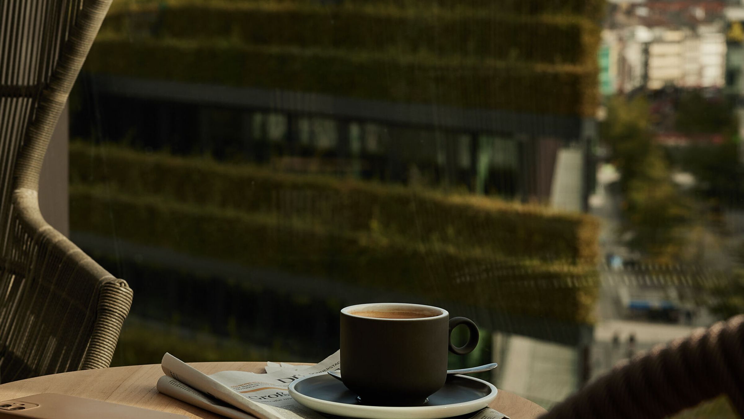 Balkon mit bequemen Stühlen und kleinem Tisch, darauf eine Kaffeetasse und eine Zeitung, mit Blick auf eine grüne Dachterrasse und die Stadt im Hintergrund. Balkon mit bequemen Stühlen und kleinem Tisch, darauf eine Kaffeetasse und eine Zeitung, mit Blick auf eine grüne Dachterrasse und die Stadt im Hintergrund.