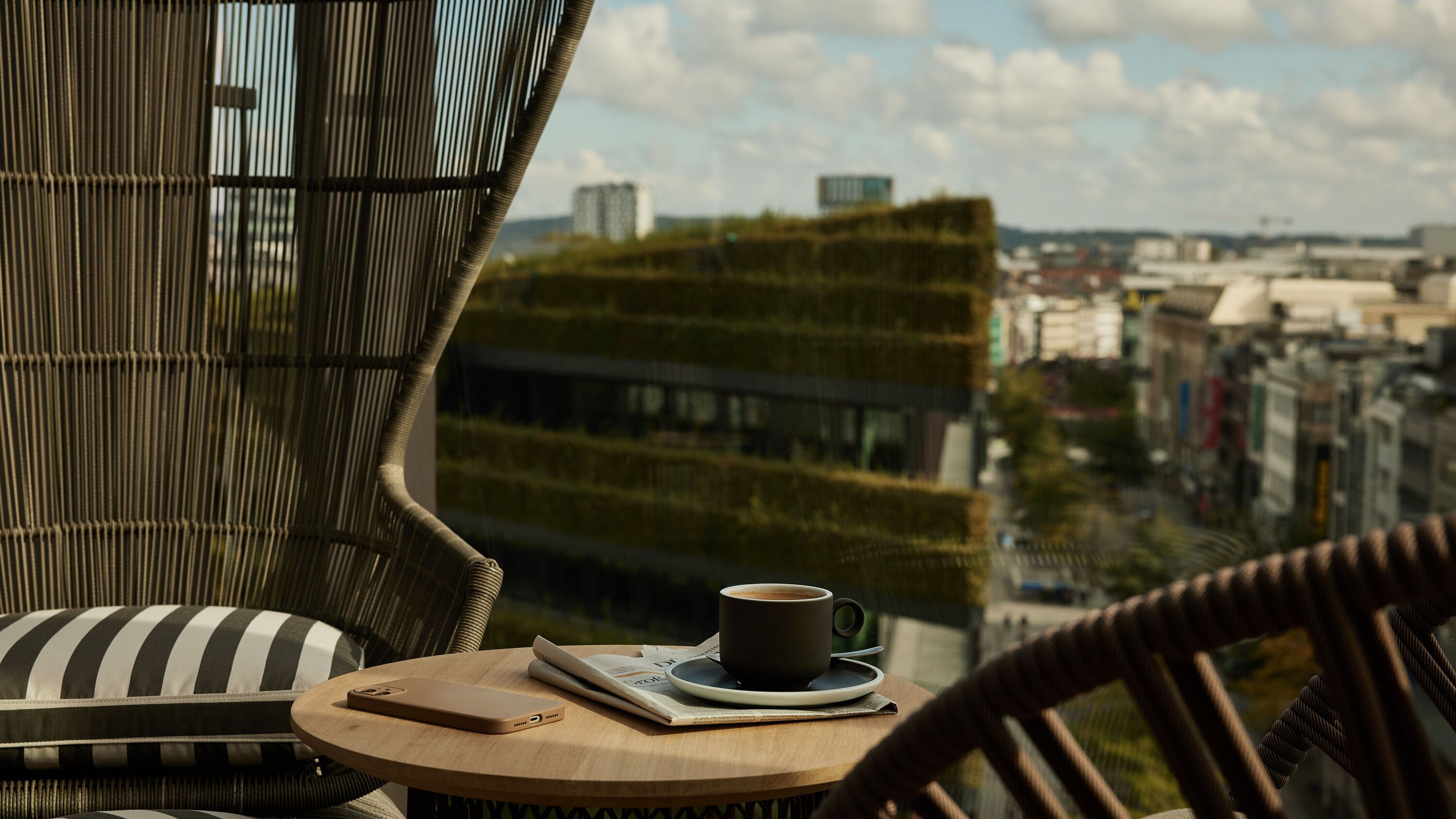 Balkon mit bequemen Stühlen und kleinem Tisch, darauf eine Kaffeetasse und eine Zeitung, mit Blick auf eine grüne Dachterrasse und die Stadt im Hintergrund.