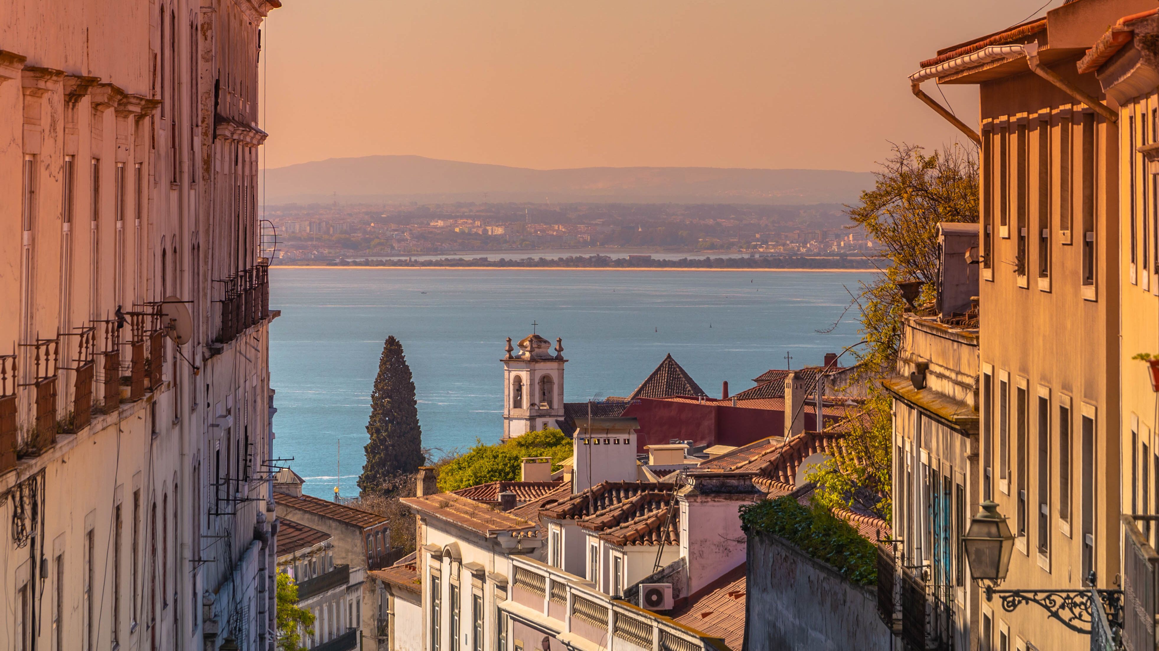 Blick durch eine enge Straße mit traditionellen Gebäuden zu einem Fluss oder Meer, im Hintergrund sanfte Hügel und ein Kirchturm bei warmem Abendlicht.