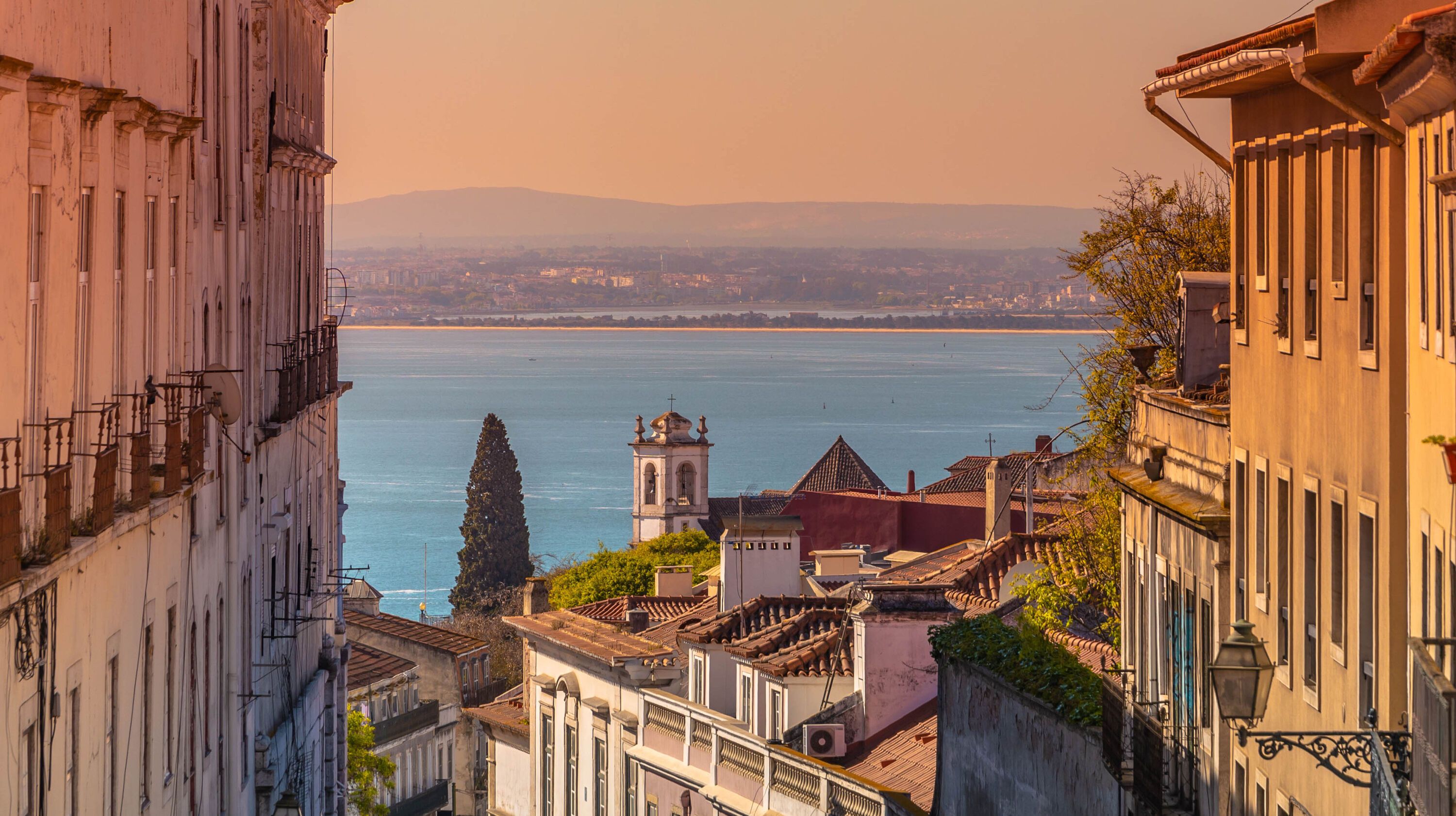 Blick durch eine enge Straße mit traditionellen Gebäuden zu einem Fluss oder Meer, im Hintergrund sanfte Hügel und ein Kirchturm bei warmem Abendlicht.