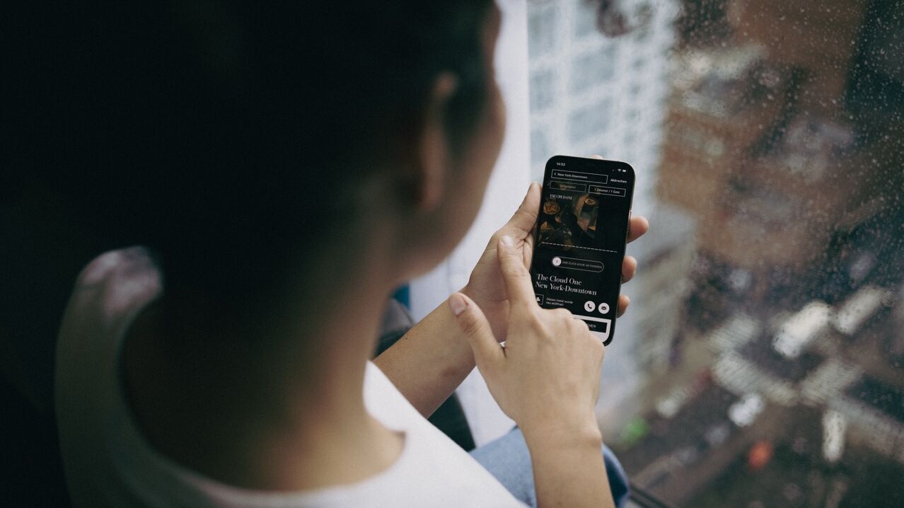 Person sitzt entspannt am Fenster und nutzt ein Smartphone, um Musik oder Podcasts zu hören, während es draußen regnet. Atmosphäre wirkt ruhig und gemütlich.