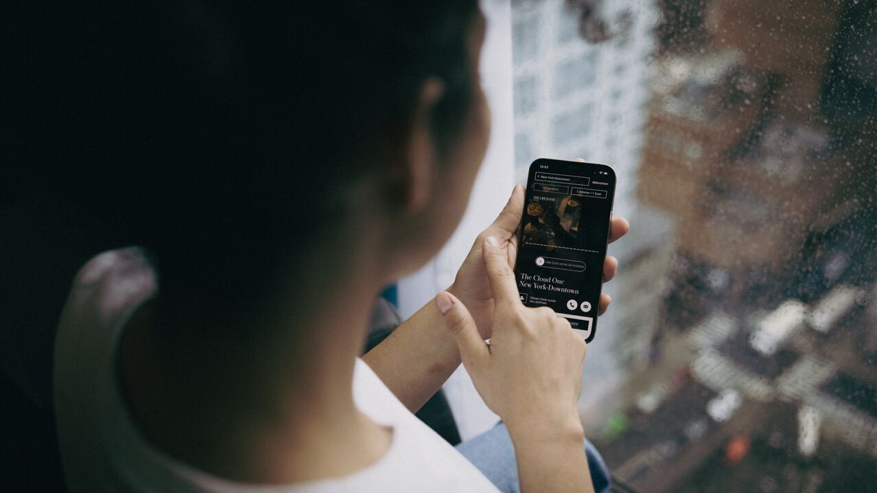 Person sitzt entspannt am Fenster und nutzt ein Smartphone, um Musik oder Podcasts zu hören, während es draußen regnet. Atmosphäre wirkt ruhig und gemütlich.