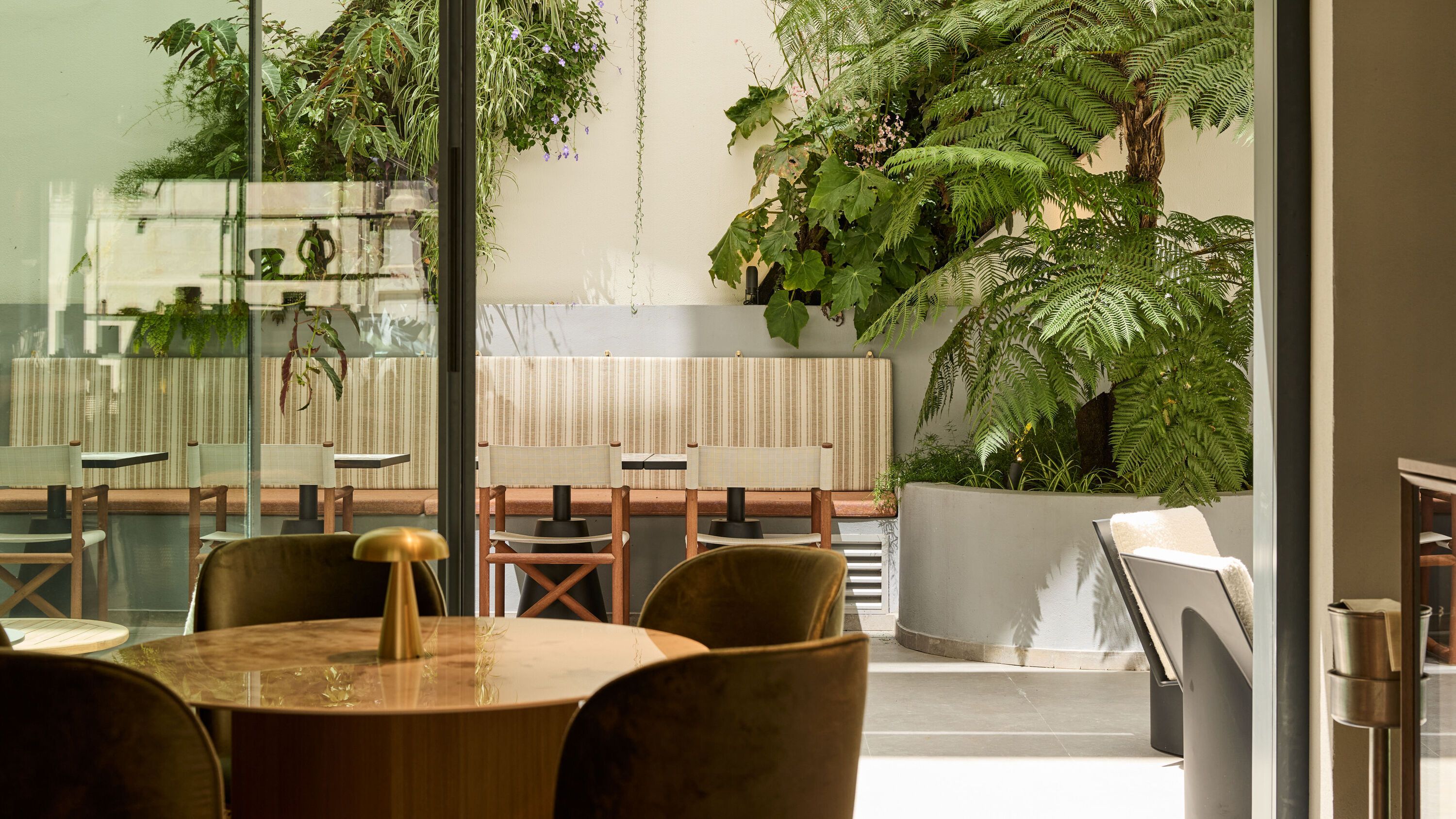 Hotel Lissabon The Cloud One Lounge-Bereich mit runden Tischen, bequemen Stühlen und großer Fensterfront, die auf eine grüne, pflanzenreiche Terrasse hinausblickt.