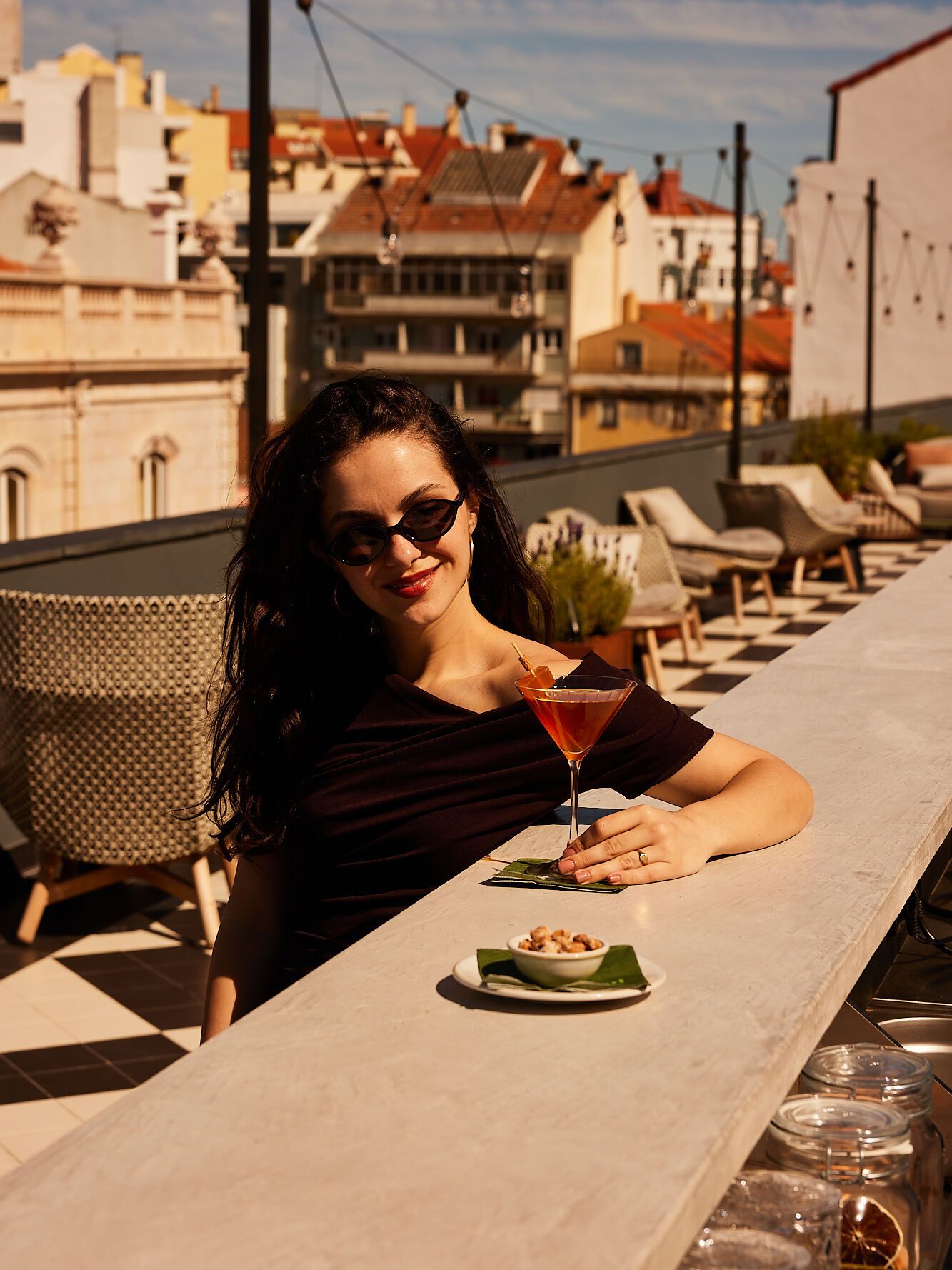 Frau sitzt entspannt auf einer Dachterrasse, genießt bei Sonnenschein einen Cocktail und eine kleine Snackschale mit Blick auf die Stadt.