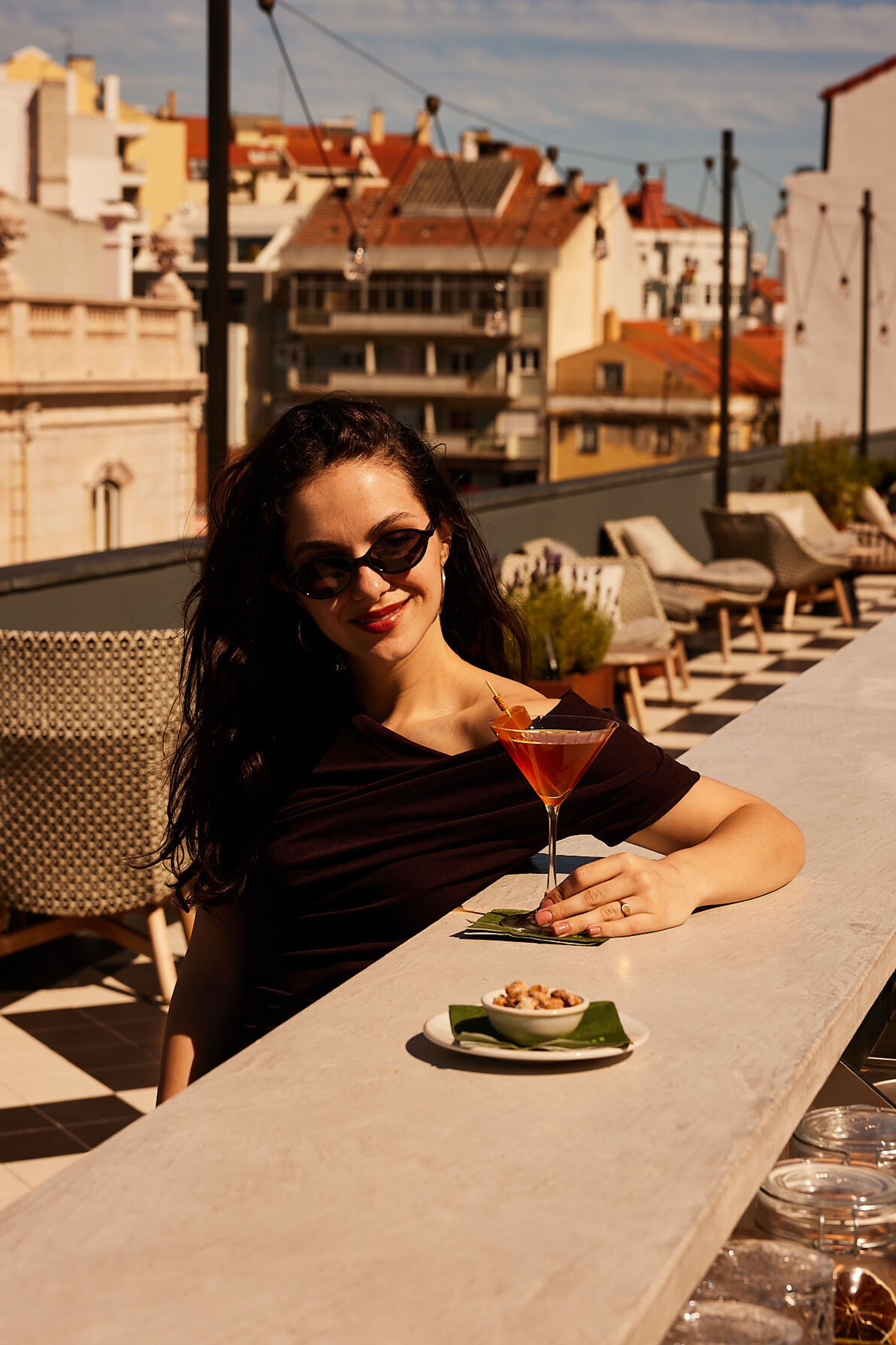 Frau sitzt entspannt auf einer Dachterrasse, genießt bei Sonnenschein einen Cocktail und eine kleine Snackschale mit Blick auf die Stadt.