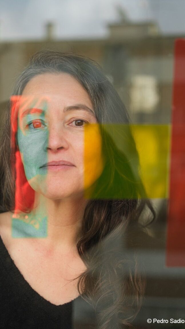 Porträt einer nachdenklichen Frau hinter einer Glasscheibe, durch die sich verschwommene bunte Farbfelder in Rot, Gelb und Grün spiegeln.