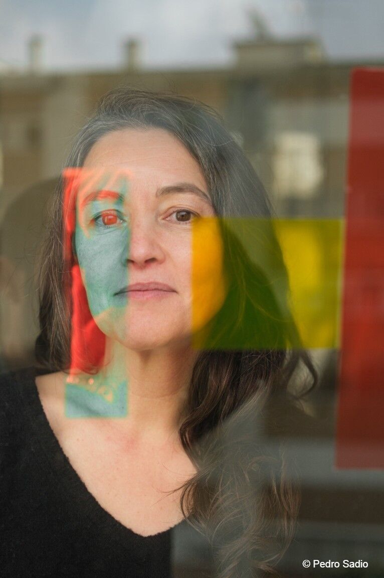 Porträt einer nachdenklichen Frau hinter einer Glasscheibe, durch die sich verschwommene bunte Farbfelder in Rot, Gelb und Grün spiegeln.