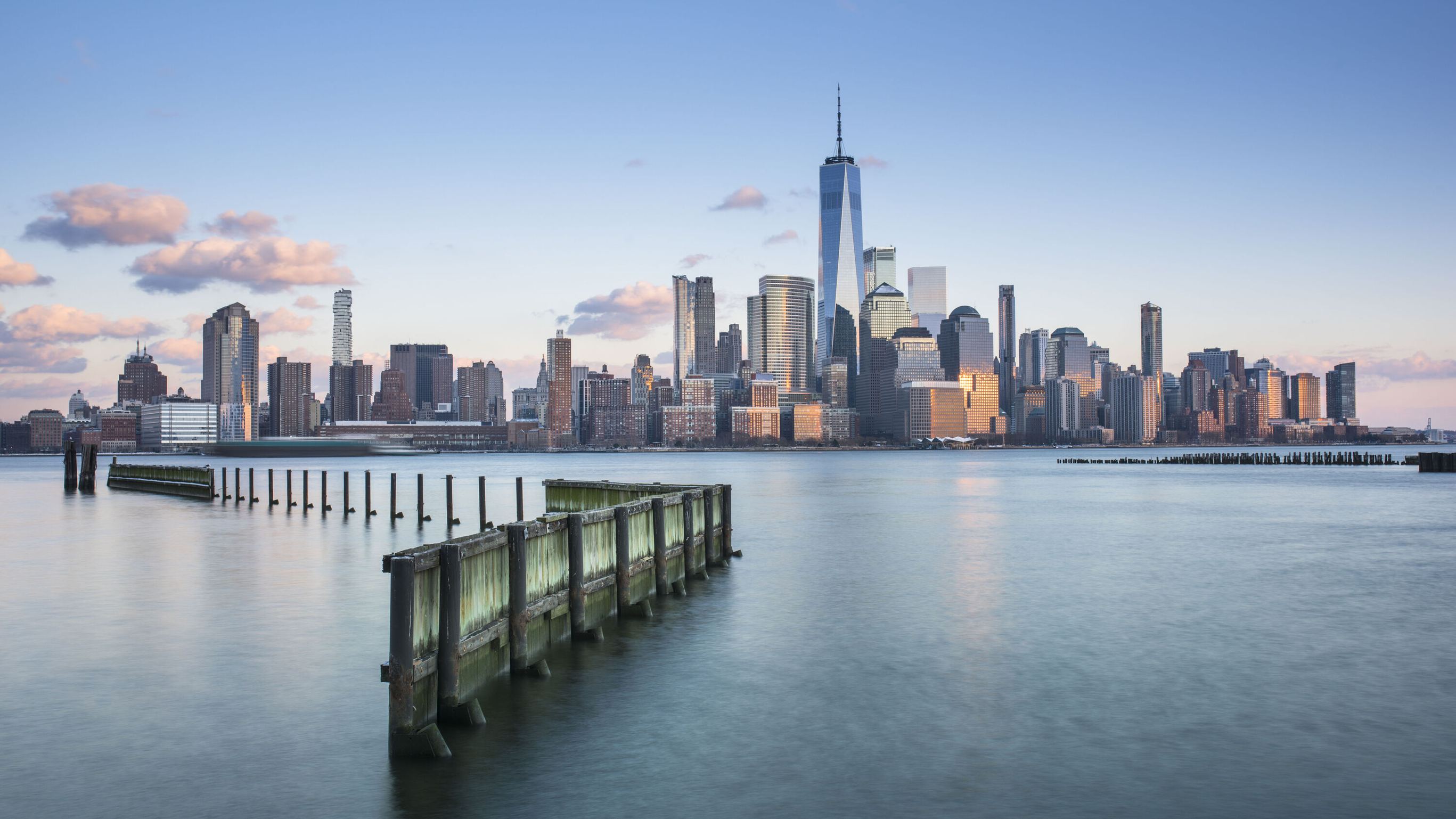 Blick auf die Skyline von Manhattan bei klarem Himmel, mit dem markanten One World Trade Center und ruhigem Wasser im Vordergrund, das eine alte Holzpier umspült.