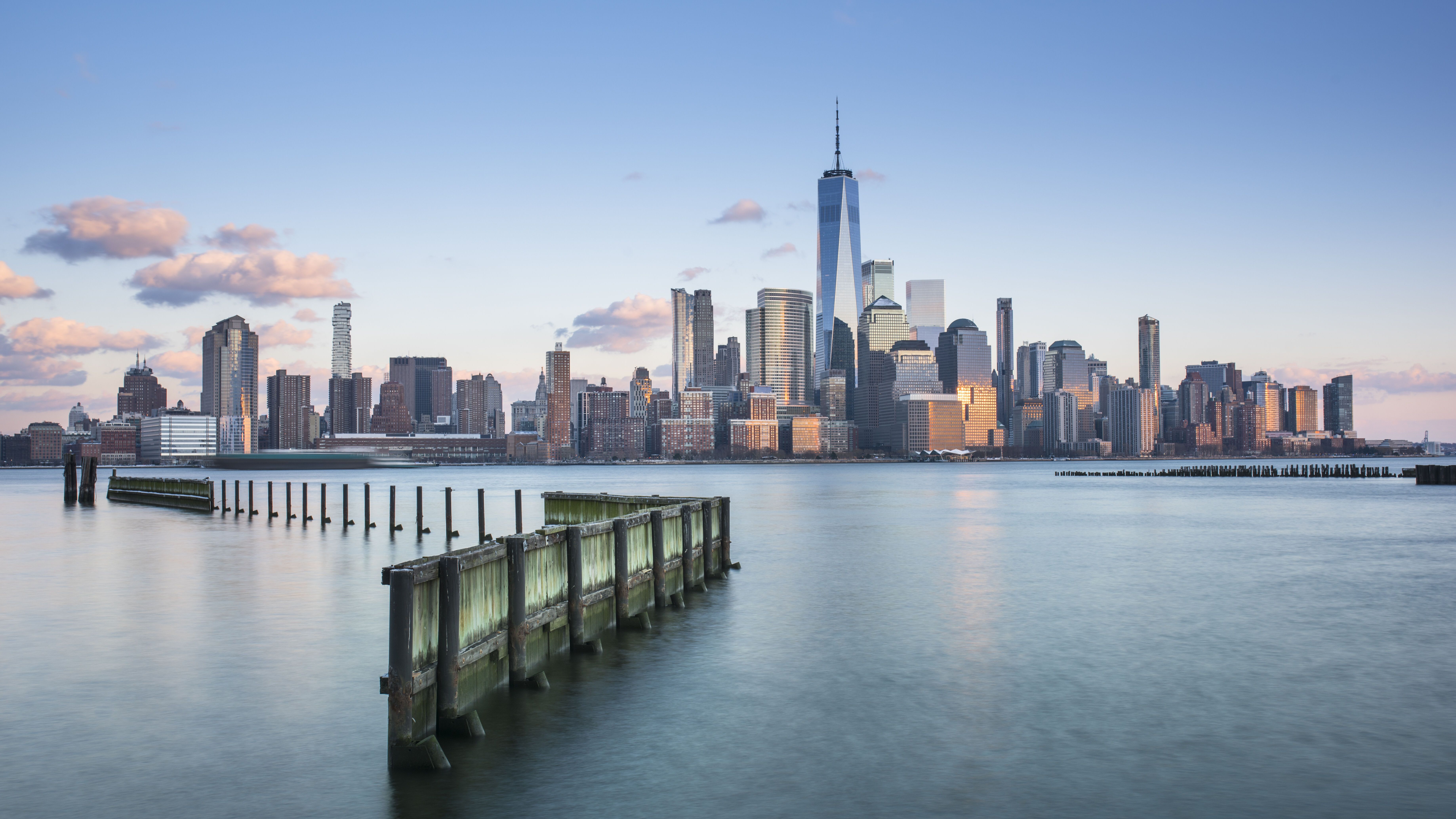 Blick auf die Skyline von Manhattan bei klarem Himmel, mit dem markanten One World Trade Center und ruhigem Wasser im Vordergrund, das eine alte Holzpier umspült.